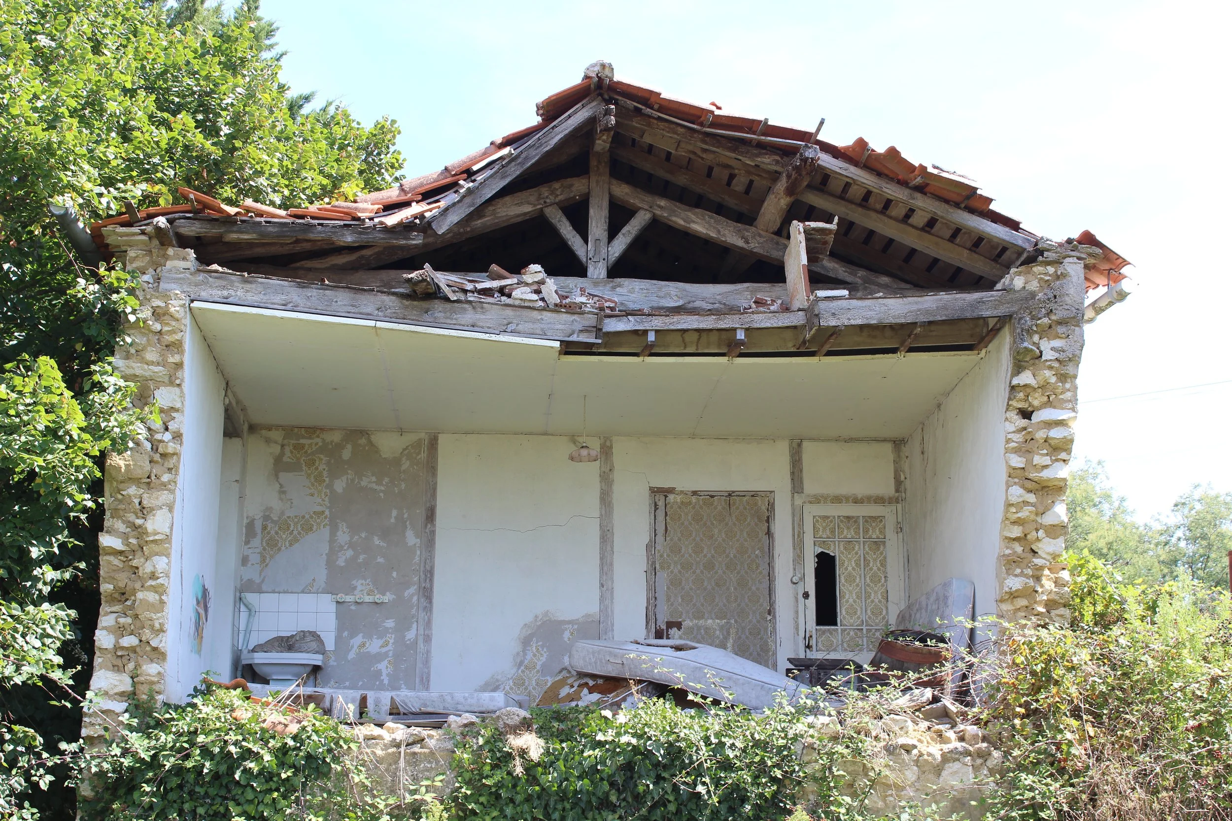 Ein verfallenes, teilweise eingestürztes Haus mit zerbrochenem Fenster und umgestürztem Sofa, umgeben von Bewuchs und Bäumen.