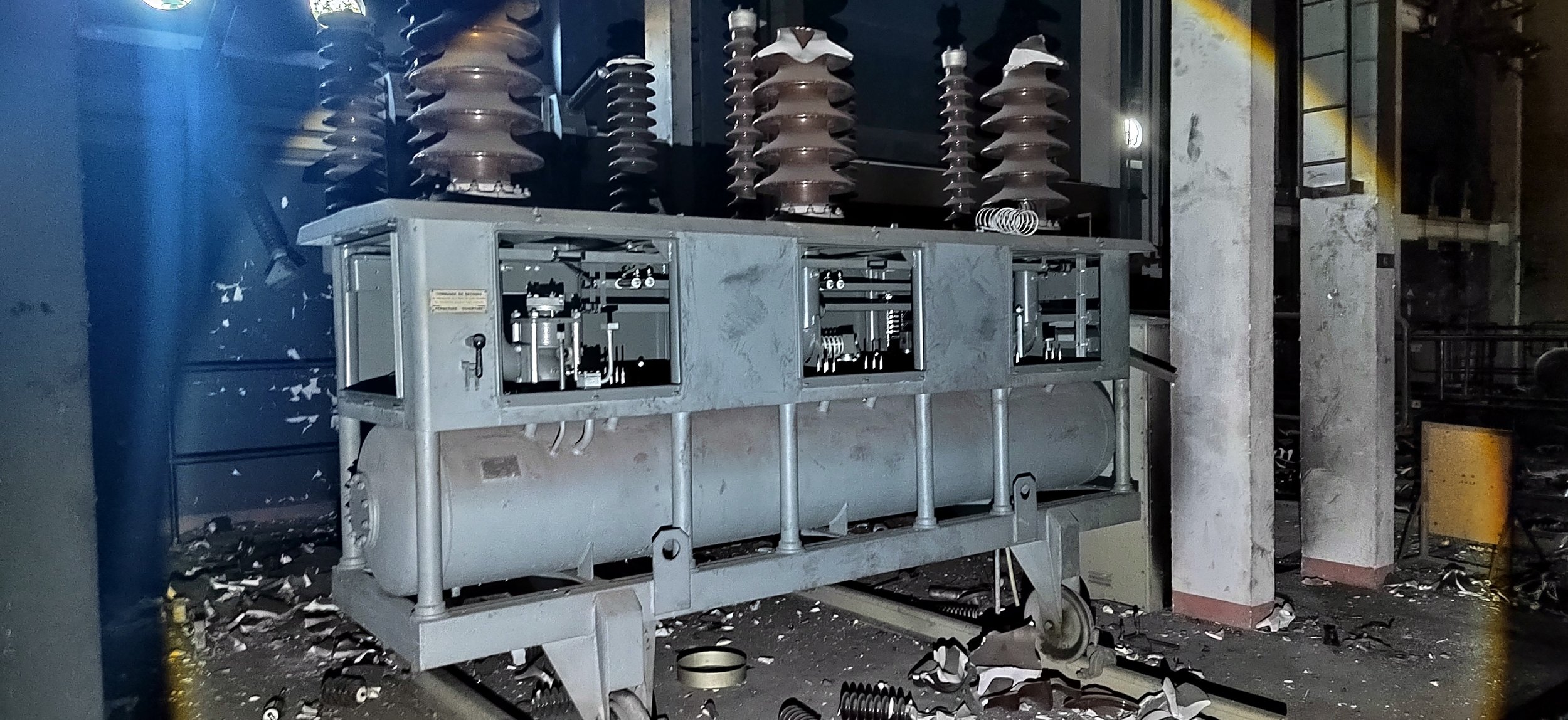 Ein defekter elektrischer Transformator mit zerstörten Isolatoren und beschädigten Komponenten in einer dunklen, verschmutzten Umgebung.