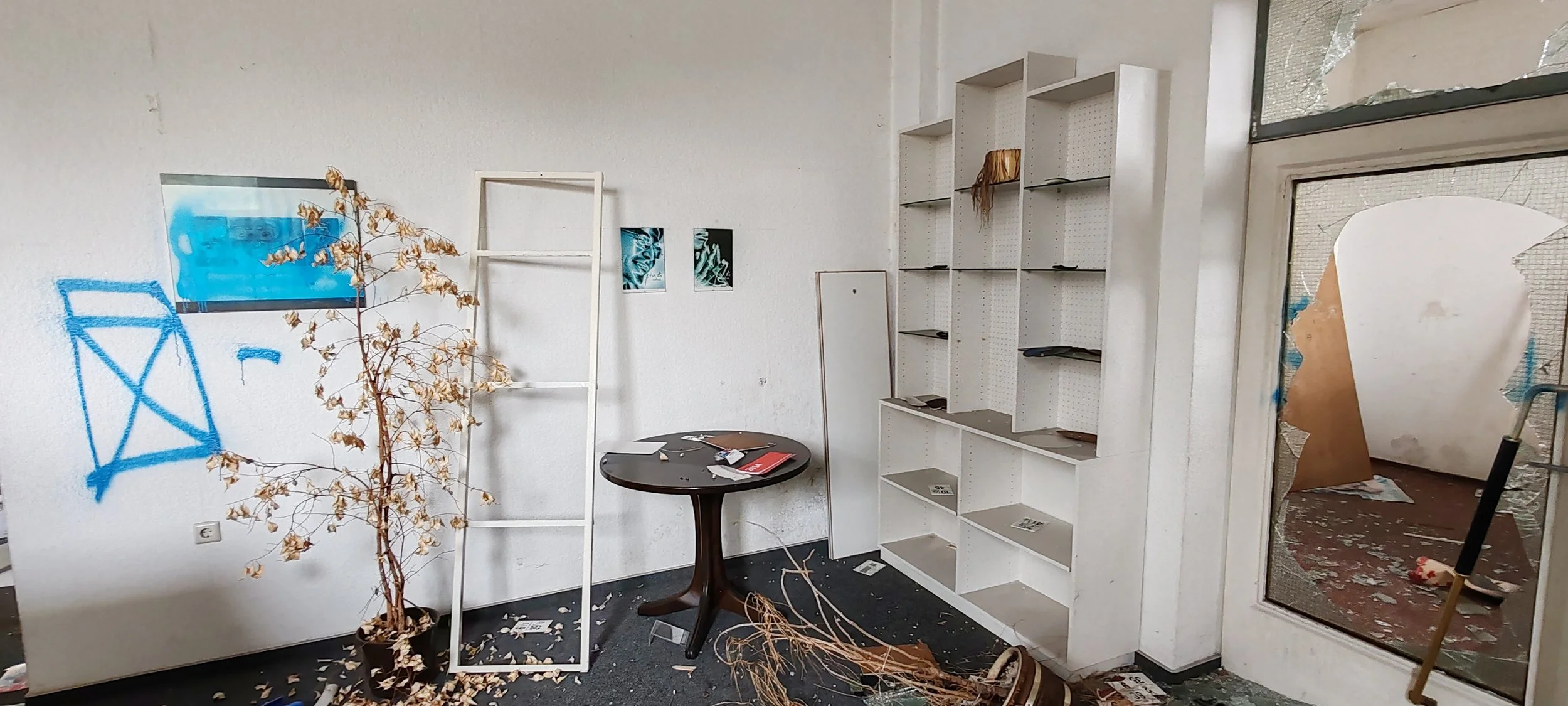 Ein verlassener Raum mit weißen Wänden, zerbrochenen Fensterscheiben, einer leeren weißen Regal, einem schwarzen Tisch mit Papieren, einem vertikalen Spiegel und einer toten Pflanze, die auf dem Boden und auf einem Ständer liegt, sowie Graffiti auf d