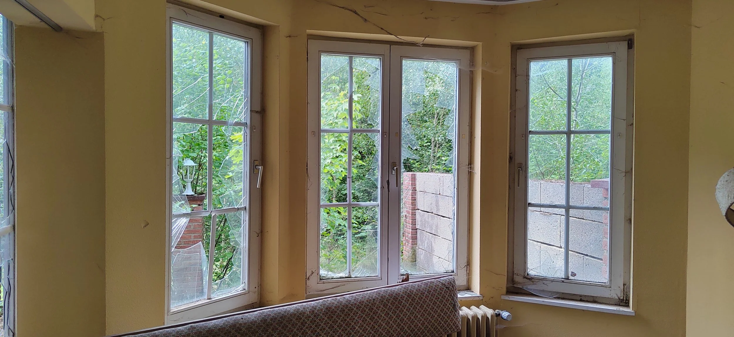 Drei große Fenster mit weißen Rahmen und Gitter, durch die man grüne Bäume und einen Zaun im Garten sehen kann, in einer gelben Wand. Das Fenster auf der linken Seite ist zerbrochen, mit zerbrochenem Glas. Es gibt einen alten Heizkörper unter den Fenstern.