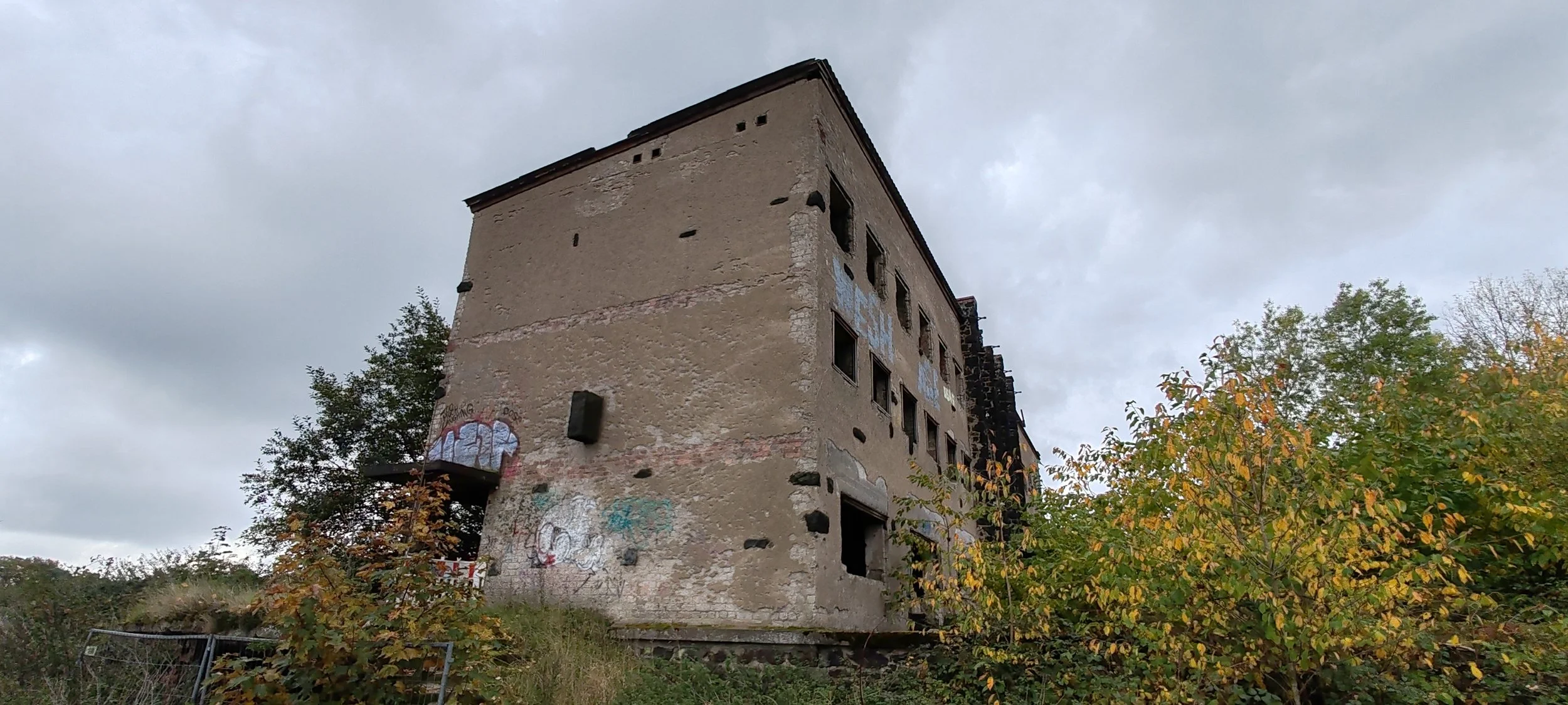 Altes verfallenes Gebäude mit Graffiti an der Wand, umgeben von Bäumen mit Herbstlaub, bei bewölktem Himmel.