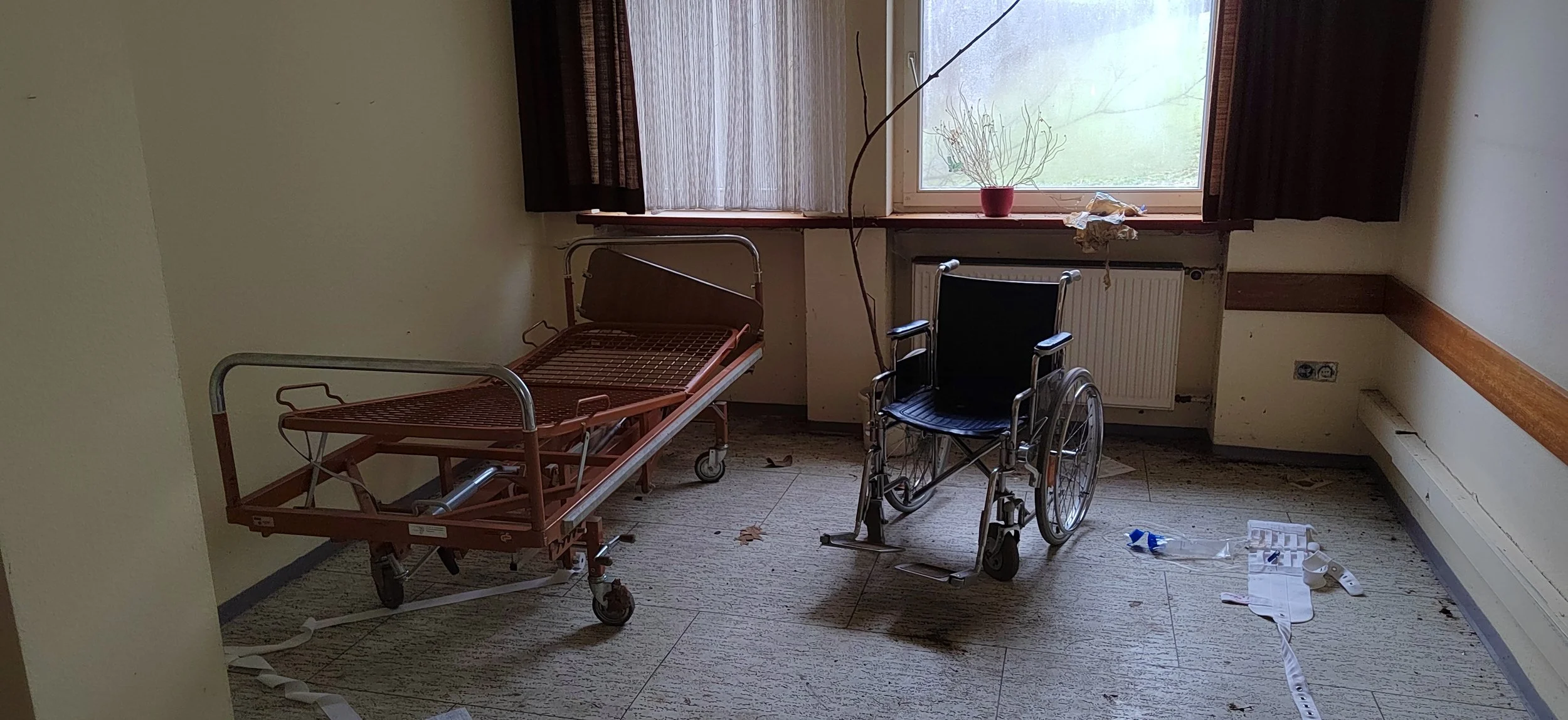 Leeres Krankenzimmer mit Krankenhaus Bett und Rollstuhl, verschmutztem Boden und zerzausten Vorhängen vor einem Fenster.