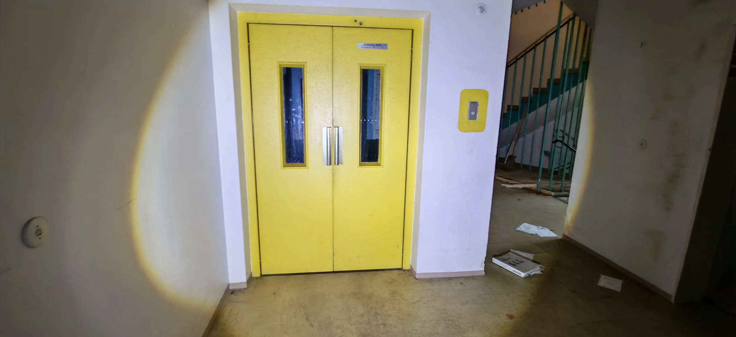 Gelber Aufzug mit Doppelglasschiebetüren und einem Digital-Display daneben, in einem Gebäude. Es gibt einen Treppenaufgang links und einige Papiere auf dem Boden.