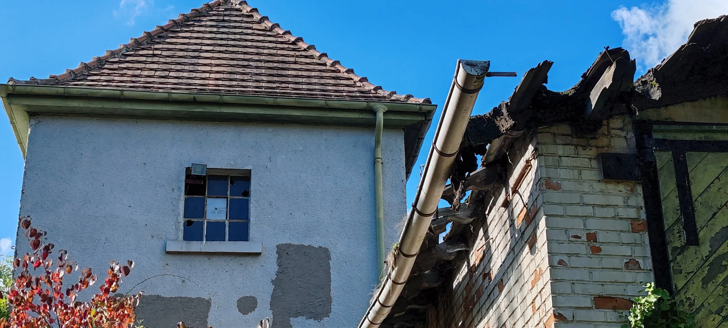 Ein beschädigtes Dach mit einer kaputten Holzkonstruktion und einer zurückgeschnittenen Mauer, daneben ein Haus mit weißverputzter Fassade und einem kleinen Fenster, unter blauem Himmel.