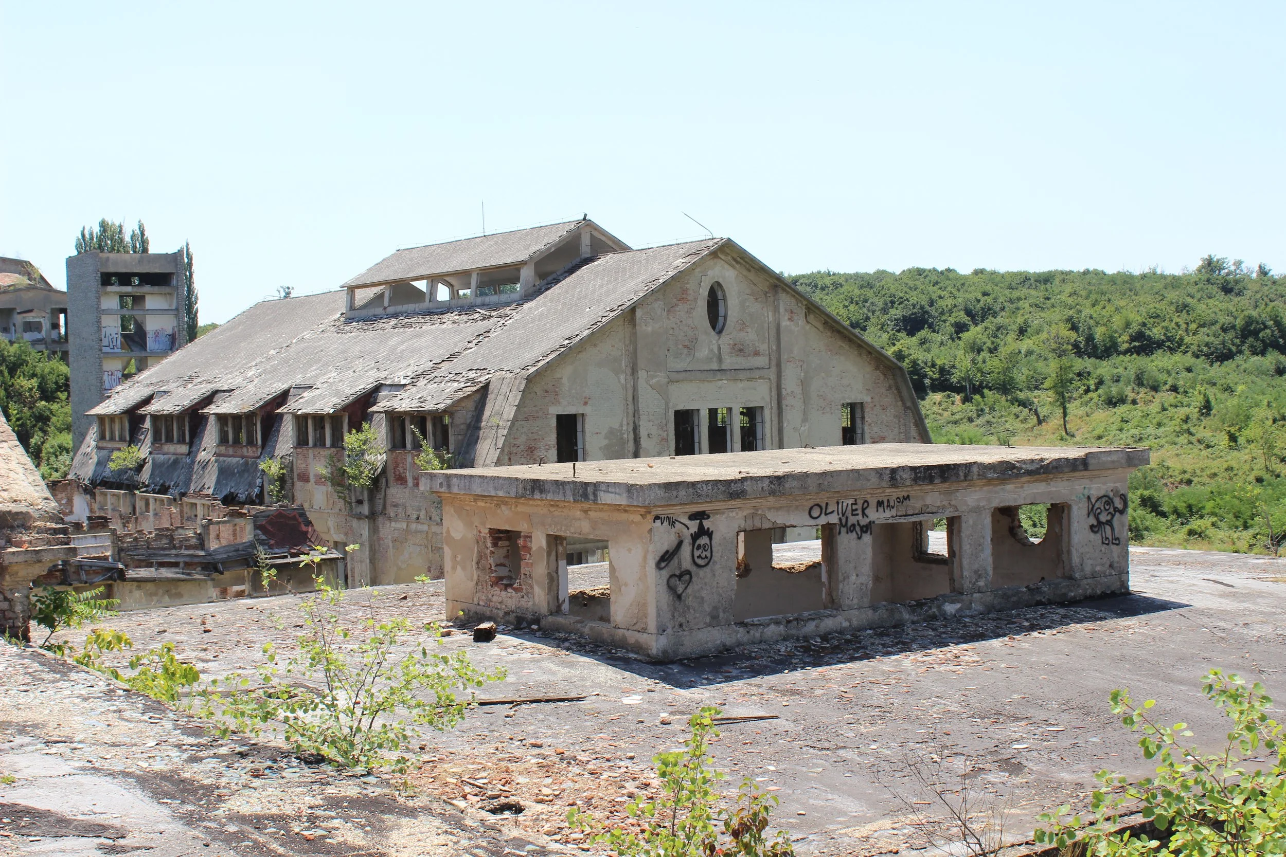 Verfallene, verlassene Gebäude auf eine unebene, zerbröckelte Fläche mit Graffiti, mit grünen Bäumen und Hügel im Hintergrund, bei sonnigem Himmel.