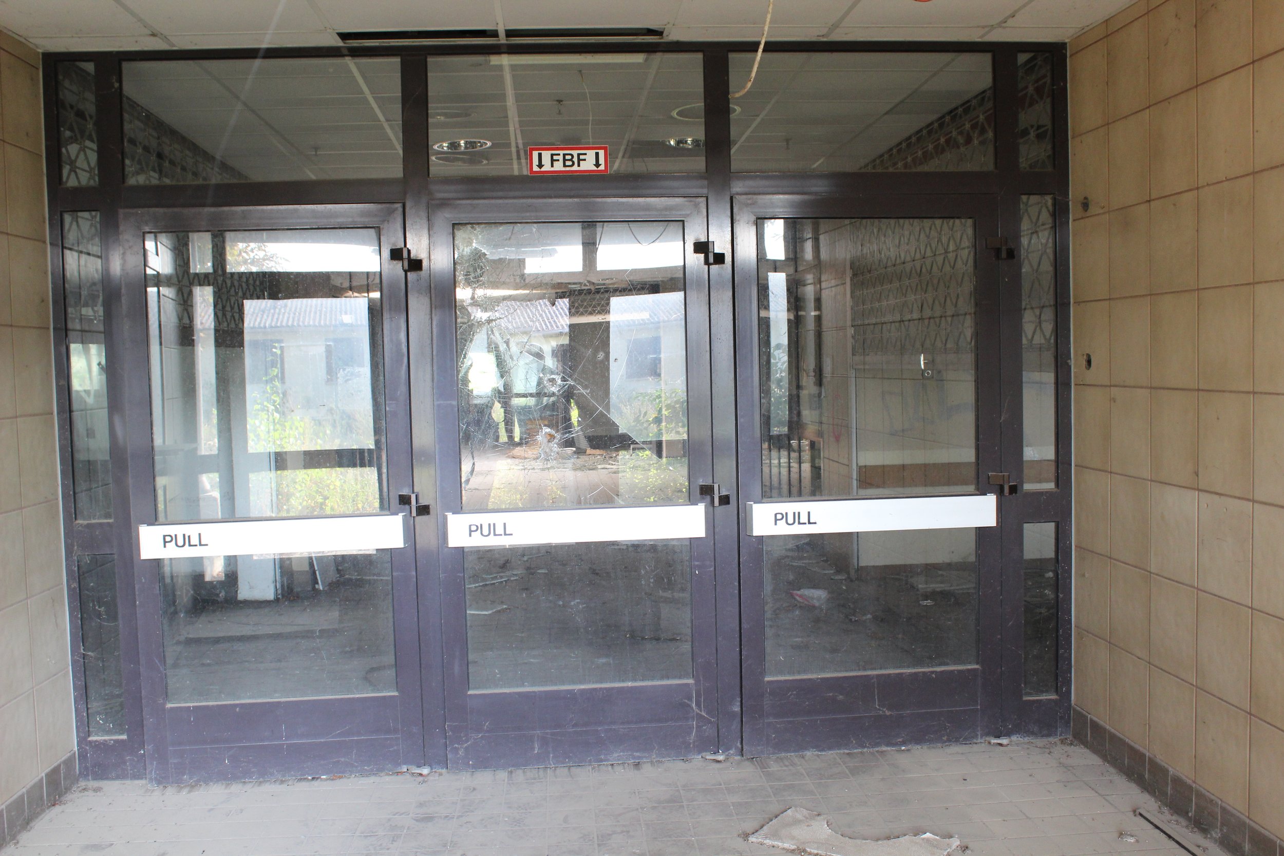 Drei doppelte Glastüren mit Metallrahmen, die im Eingangsbereich eines Gebäudes befinden. Die Türen sind schmutzig und eine ist kaputt, das Glas ist zerbrochen. Es gibt Schilder mit der Aufschrift "PULL" auf den Türen.