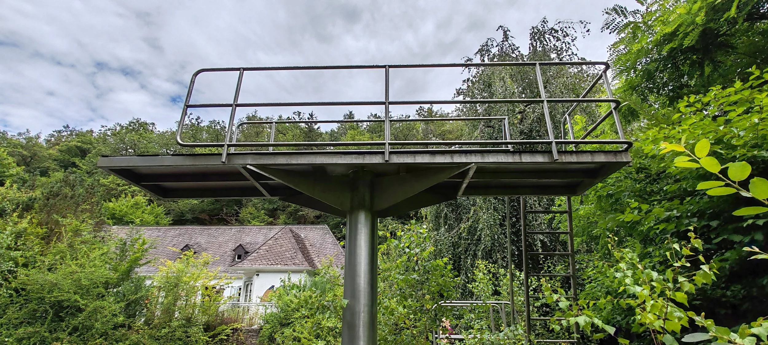 Metalner Terrasse mit Geländer, im Hintergrund ein Haus mit grauem Dach, umgeben von grünen Bäumen, bewölkter Himmel.