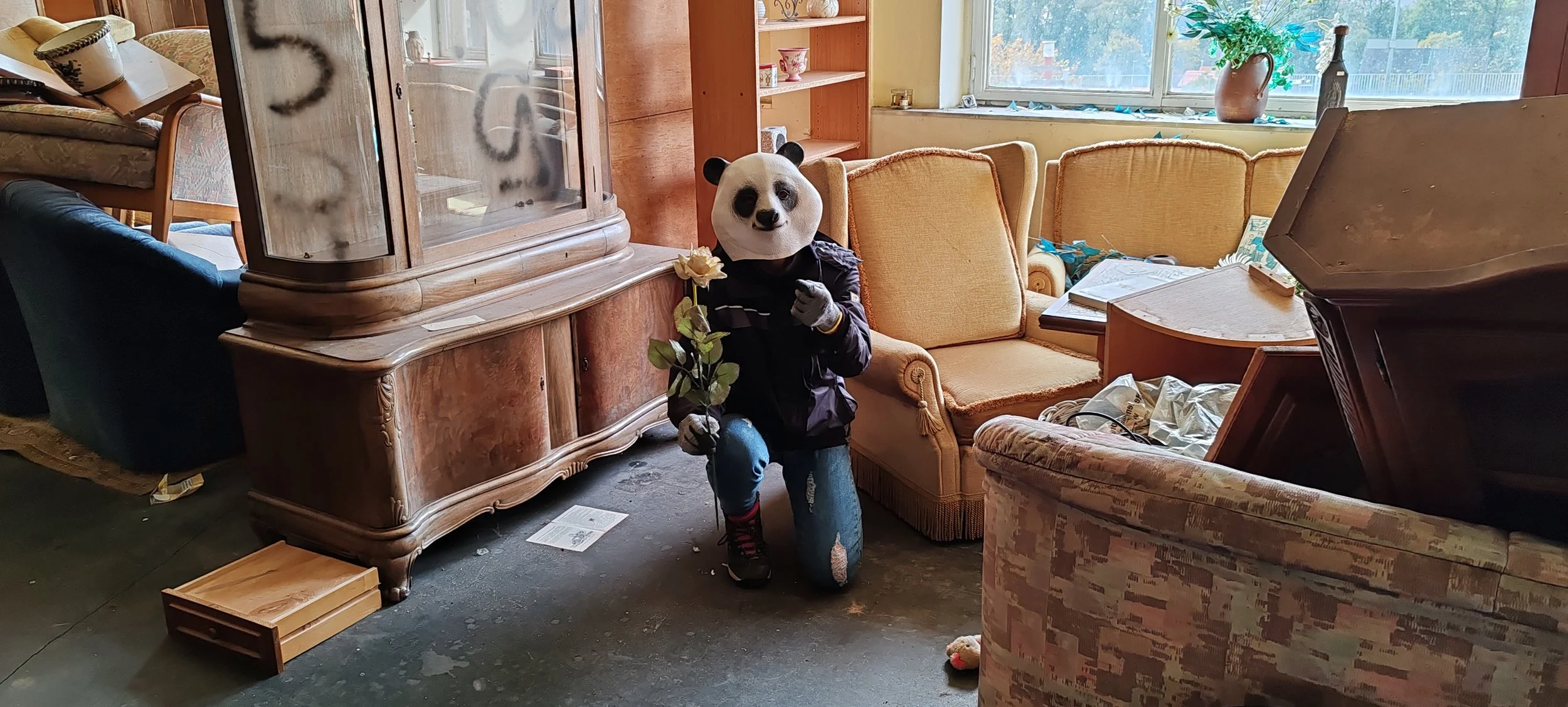 Person mit Panda-Maske, die eine große Blume hält, in einem Wohnzimmer mit alten Möbeln und großen Fenstern im Hintergrund.