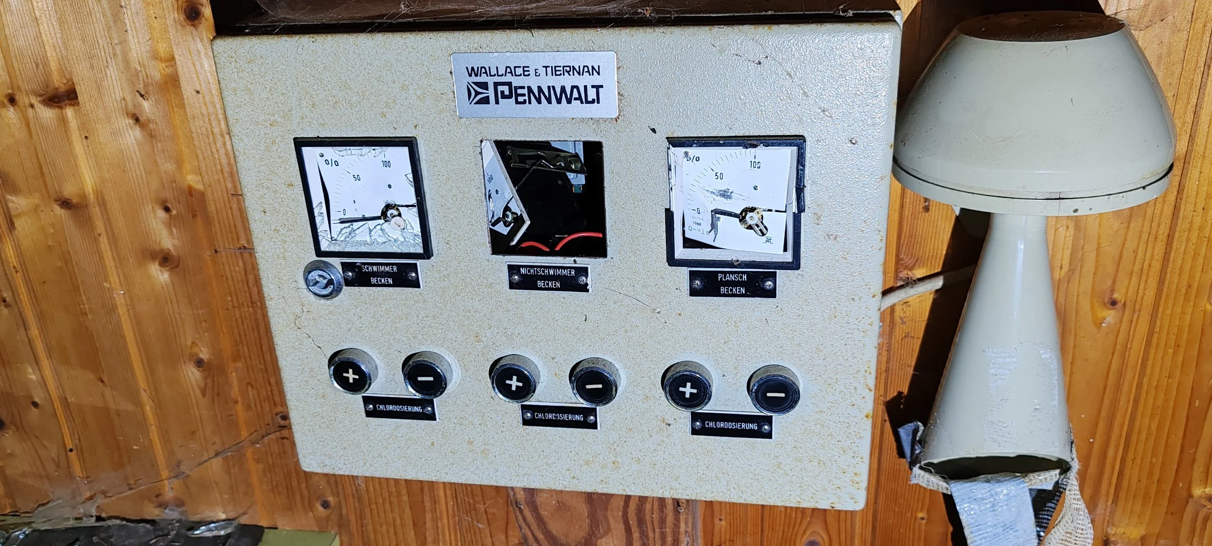 Alte Kontrolltafel mit drei Uhren und Schaltern, an einer Holzwand, mit Bezeichnungen für Becken und Chlorierung.