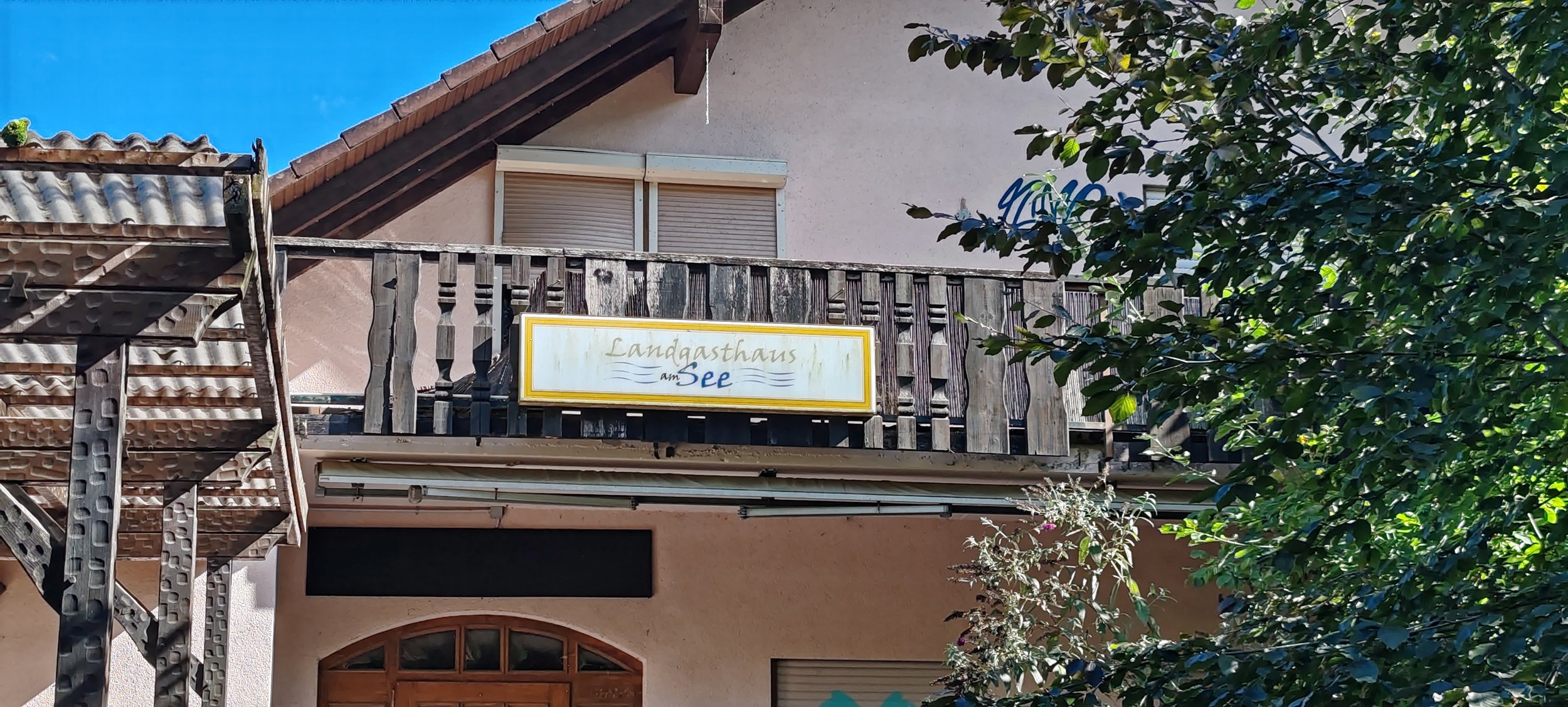 Ein Hotel mit einem Balkon, auf dem ein Schild mit der Aufschrift 'Landgasthaus am See' hängt, und davor Bäume und Äste.
