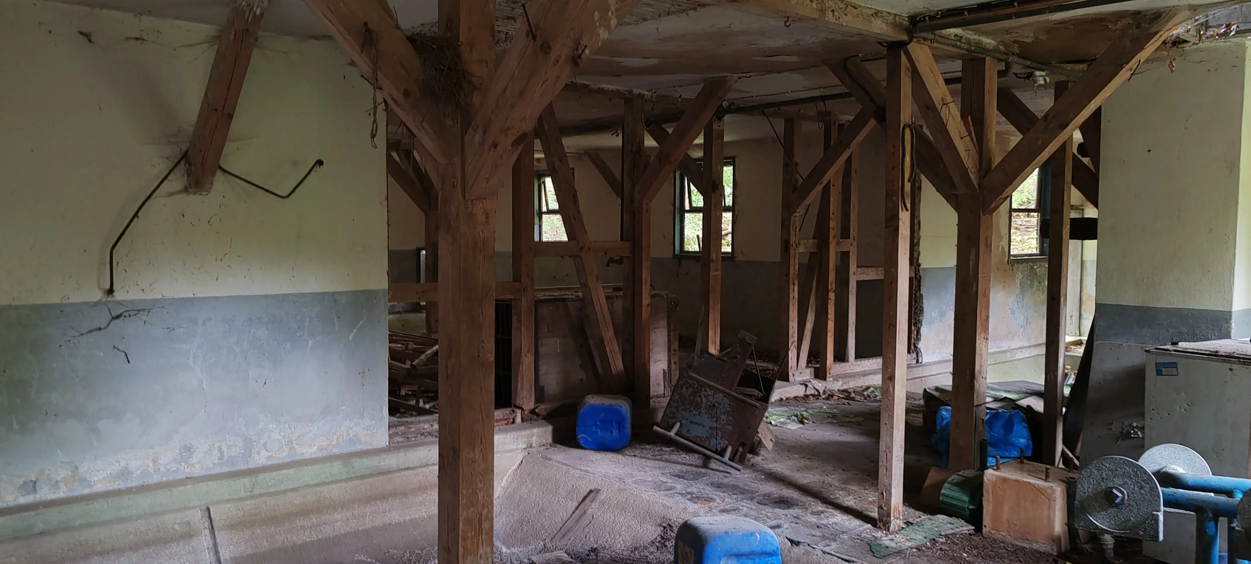 Innenansicht eines alten, unrenovierten Gebäudes mit sichtbaren Holzrahmen, Staub, alten Fenstern und verschiedenen Gegenständen im Raum.