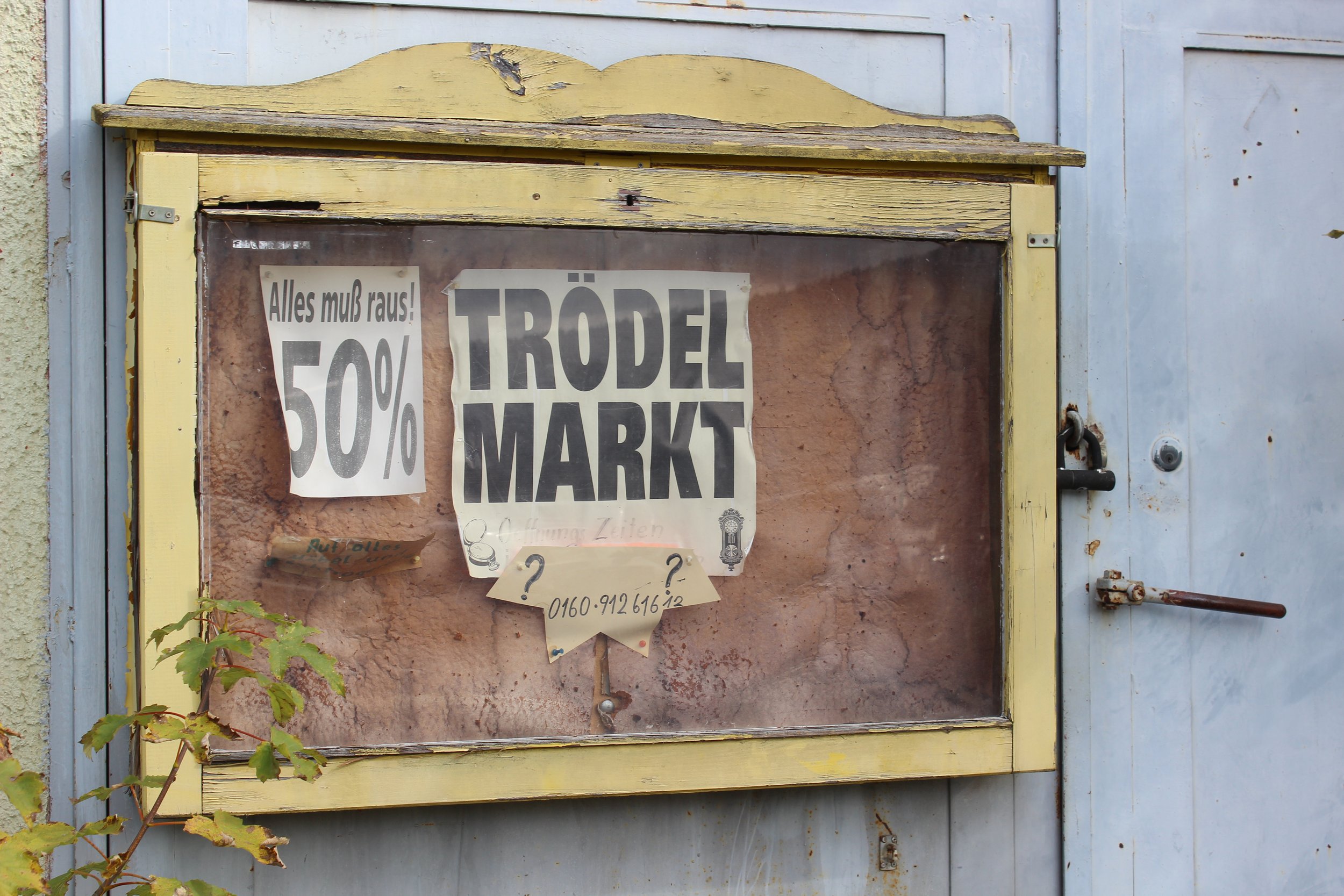 Verwitterte Holzwand mit gelber Rahmen und einem alten Glasschaukasten, der mit Postern bedeckt ist. Die Poster bewerben einen Flohmarkt mit 50% Rabatt.