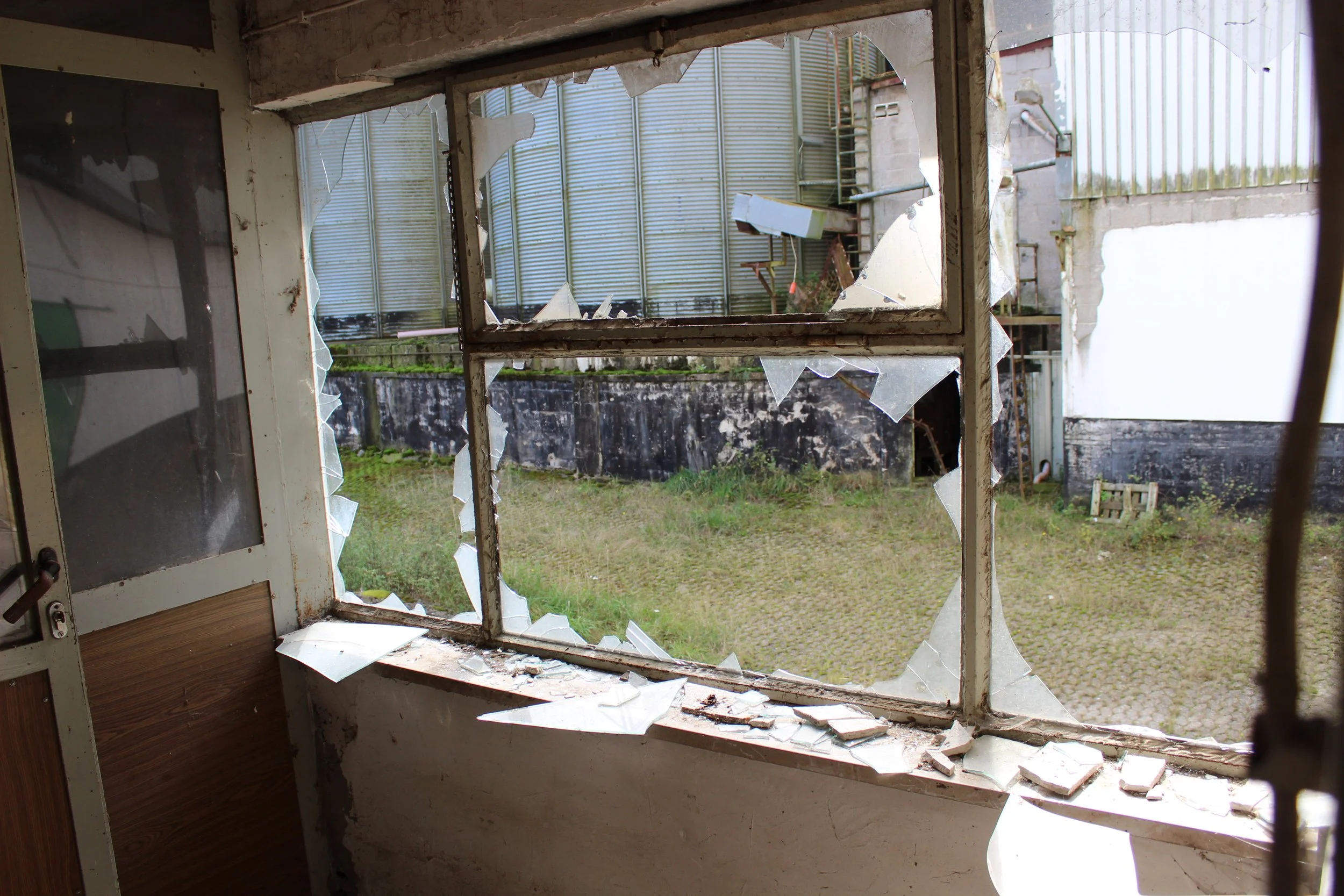 Beschädigtes Fenster mit zerbrochenem Glas, Blick nach draußen auf einen Hof mit Gras, Mauer und Lagerhallen im Hintergrund.