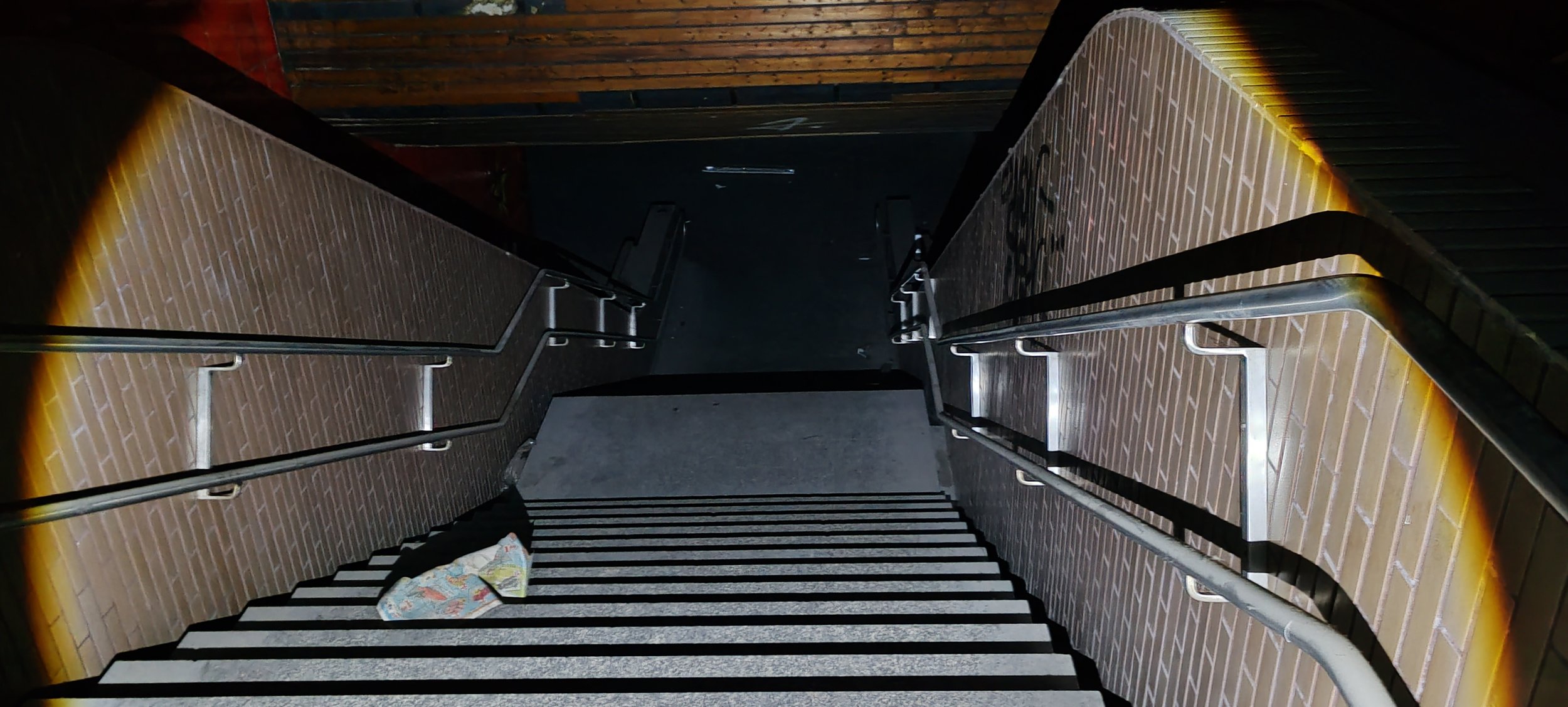 Ein Blick nach unten auf eine Treppe mit schwarzen und weißen Streifen, umgeben von Ziegelwänden mit gelblichen Beleuchtungsstreifen an den Rändern. Auf den Treppen liegt eine zerknüllte Landkarte.