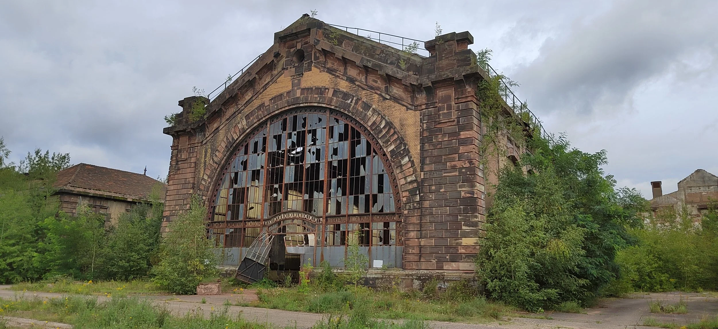 Verfallene Backsteinhalle mit zerbrochenem Fenster, überwuchert von Grünpflanzen, unter dunklem Himmel.