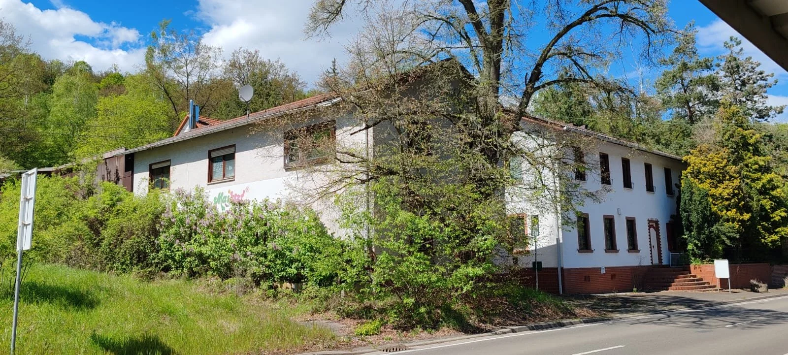 Ein weißes zweistöckiges Gebäude mit vielen Fenstern, umgeben von Bäumen und Sträuchern, bei sonnigem Wetter.