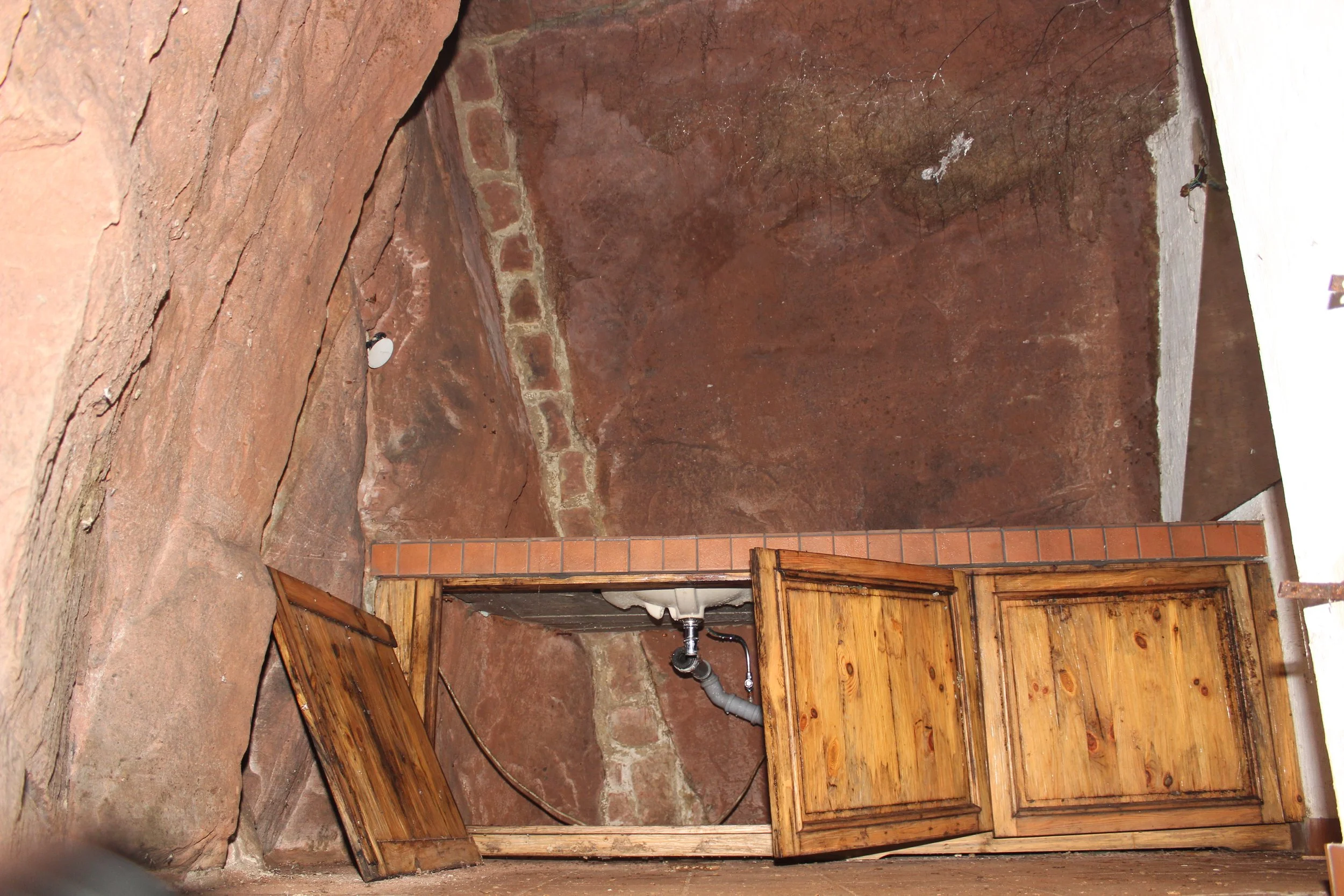 Innenbereich eines alten, geschichteten Steingewölbes mit Holzschränken, einer Wasserleitung und einer Wand aus roten Steinen.