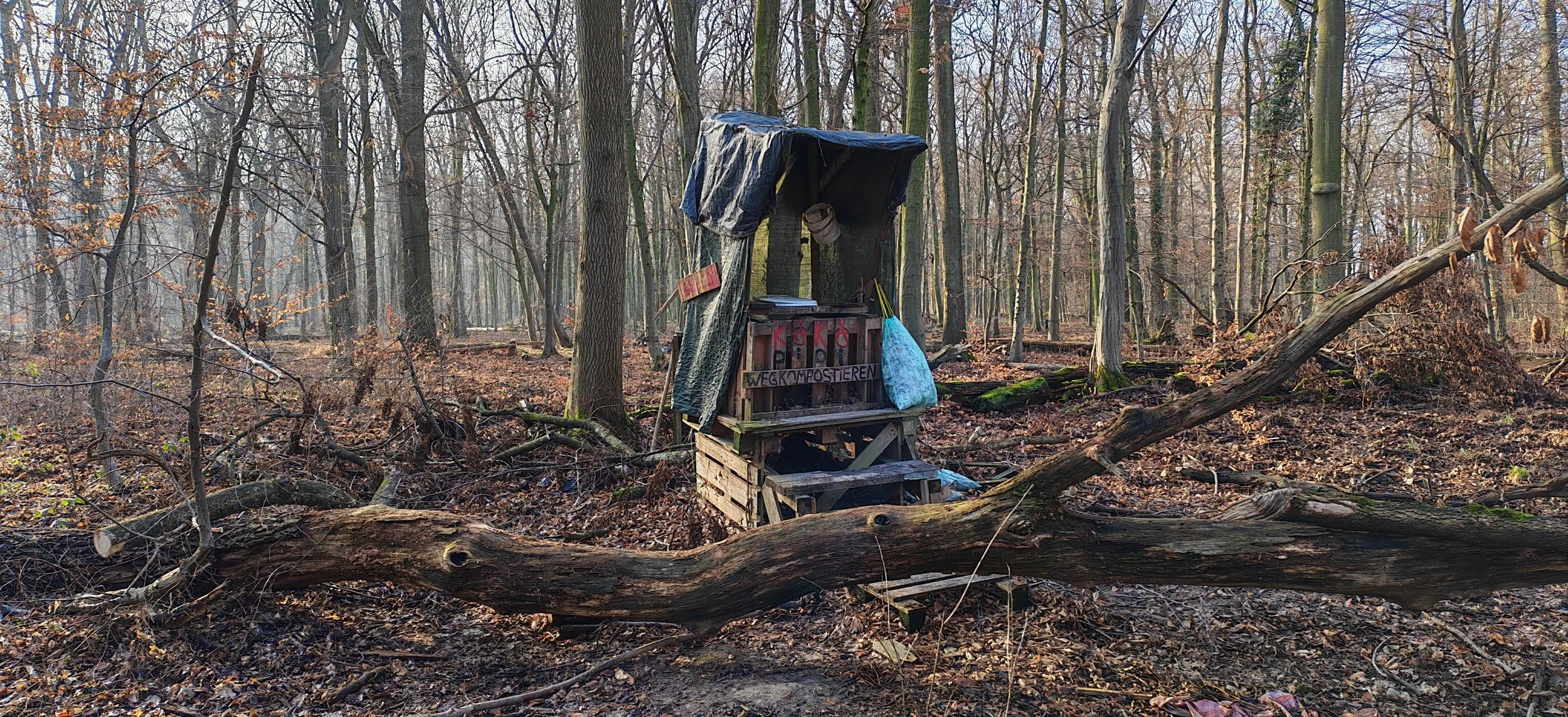 Ein improvisierter Mülleimer auf einem Holzkorpus im Wald, mit einem Schild "Weg Kompostieren" darunter, umgeben von Blätter und Bäumen.