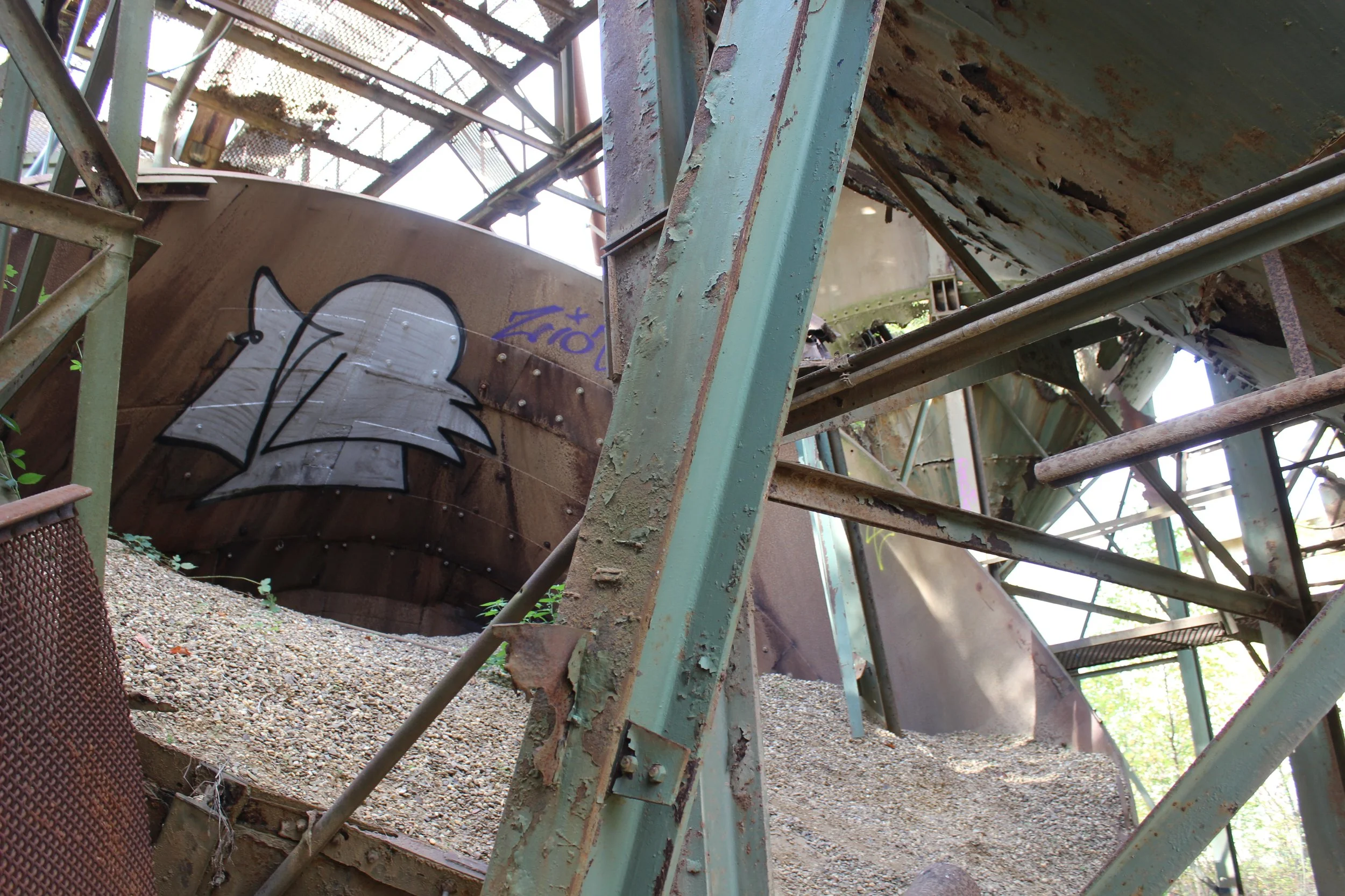 Verfallene, rostige Industrieanlage mit Graffiti auf Metallrohr, umgeben von Metallstrukturen und Sand.