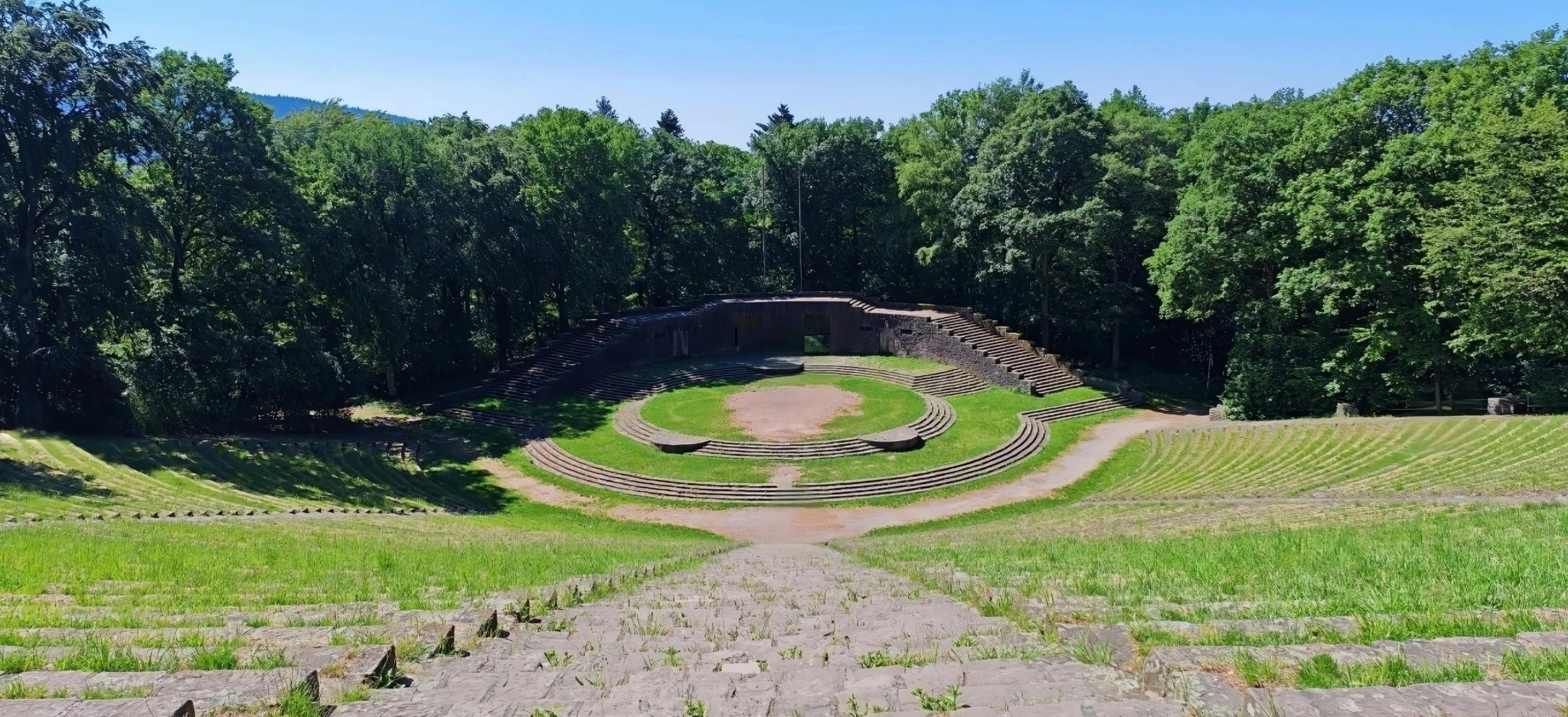 Ein verfallenes Freilichttheater im Grünen, umgeben von Bäumen, mit halb verfallener steinerner Bühne und aufgestellten Sitzreihen.