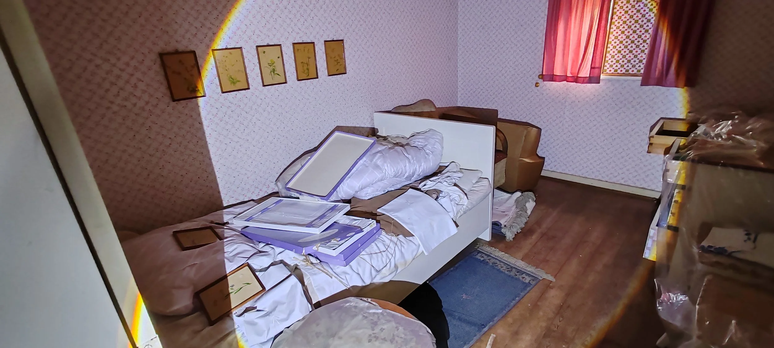 Chaos im Schlafzimmer mit aufgerissenem Bett, verstreuter Bettwäsche, Pappkartons auf dem Bett, an der Wand hängen Bilder, Fenster mit roten Vorhängen und Holzpaneele, Stauraum mit Paketen