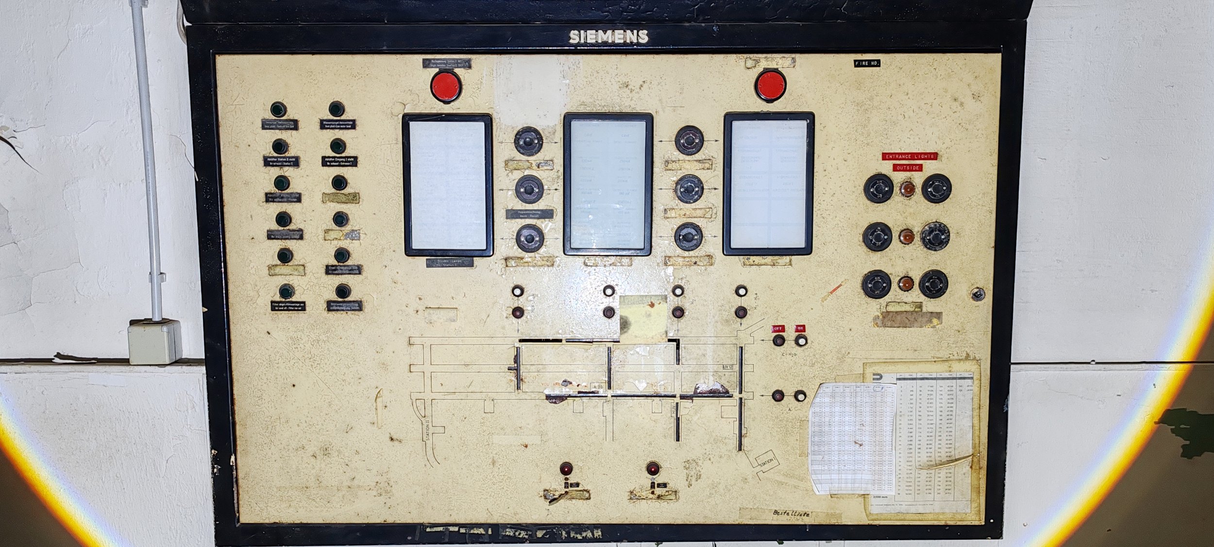 Altes, verdrecktes Steuerungspanel mit digitalen Displays, Knöpfen und Schaltern, an einer weißen Wand angebracht.