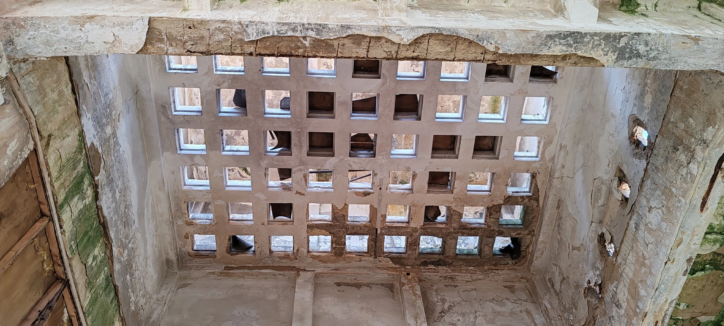 Ein verfallenes, altes Gebäude mit einer geräumigen, offenen Decke, die durch quadratische Öffnungen und teilweise zerbröckelnde Wände gekennzeichnet ist.