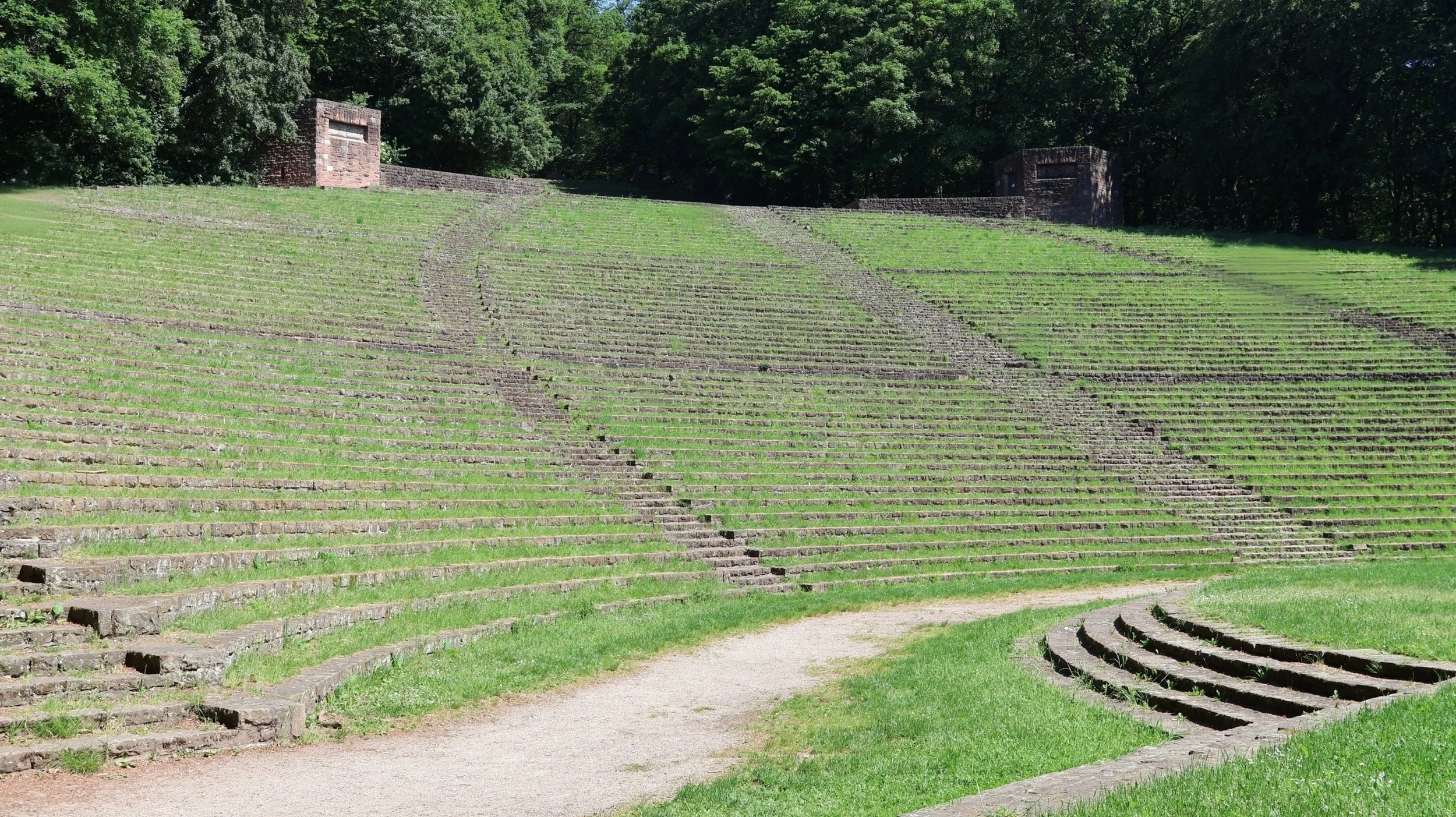 Antikes Amphitheater im Grünen mit Steinstufen und umliegenden Bäumen.