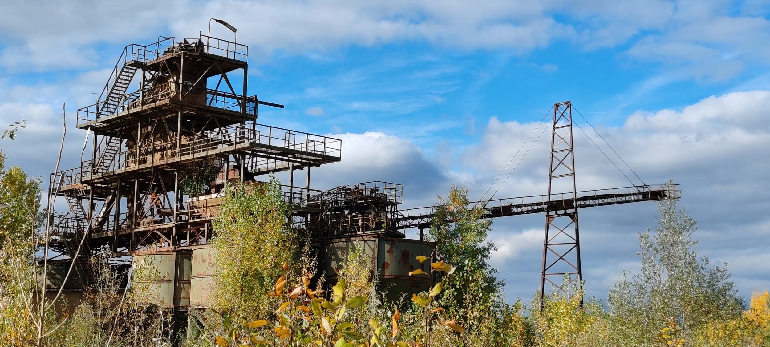 Verrostete Industrieanlage im Freien, umgeben von Büschen und Bäumen, mit blauer Himmel und Wolken im Hintergrund.
