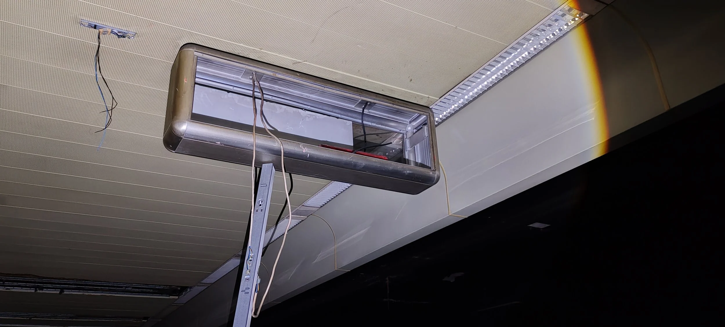 Hängendes, offengelegtes Leuchtmittel im Raum, mit sichtbaren Kabeln, an einer Leiter lehnt in einem Raum unter Deckenverkleidung.