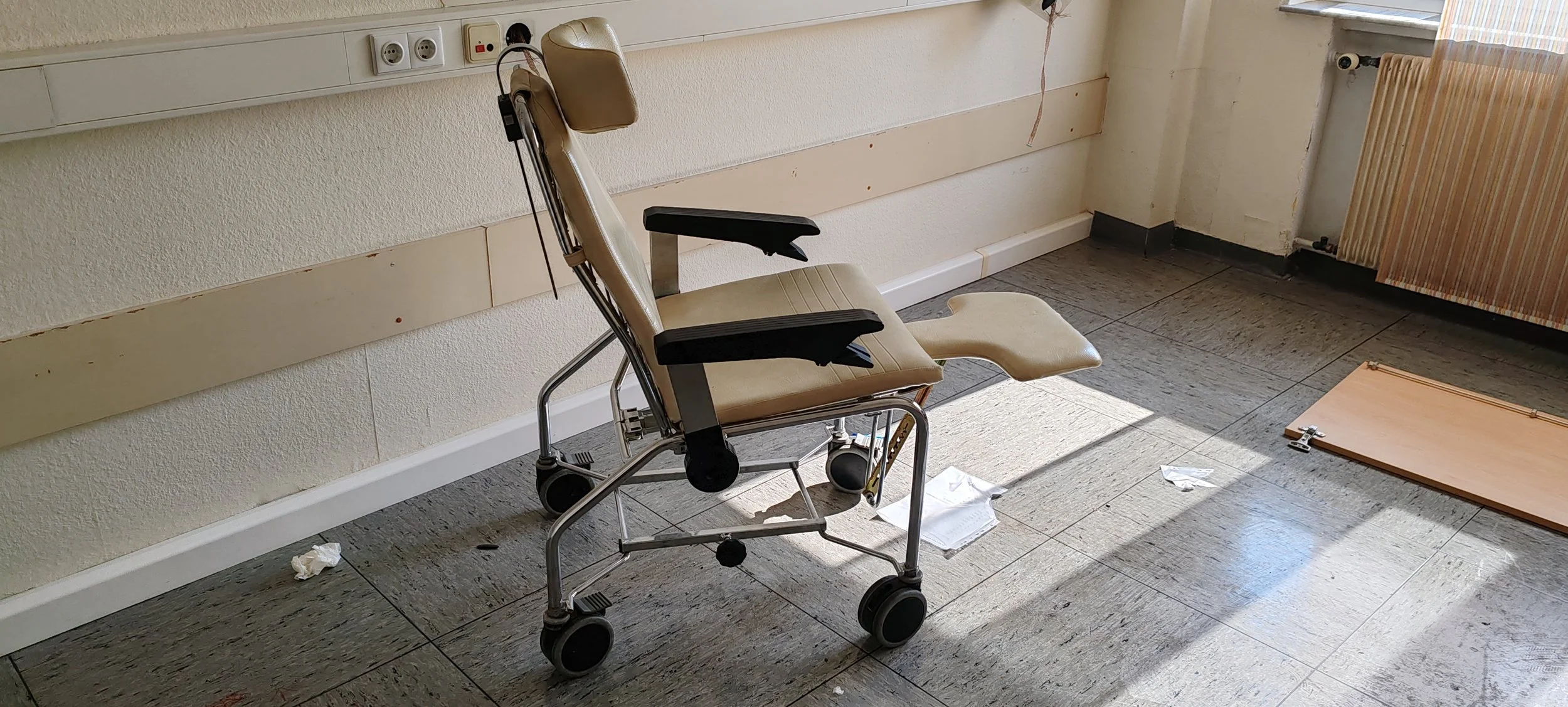 Ein veralteter Krankenhaus- oder Pflegebettstuhl in einem leeren Krankenzimmer, auf dem Boden liegen Papierstücke und ein Klappbrett.