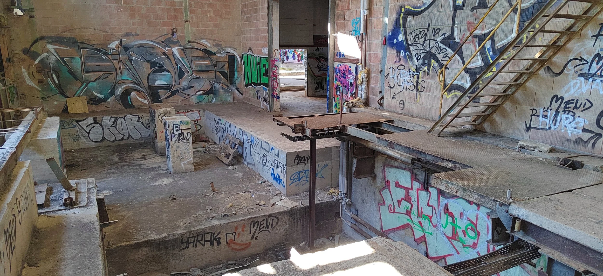 Verfallene, graffitibeschmierte leere Schwimmhalle mit offener Decke, Treppen und Wasserpfützen auf dem Boden.