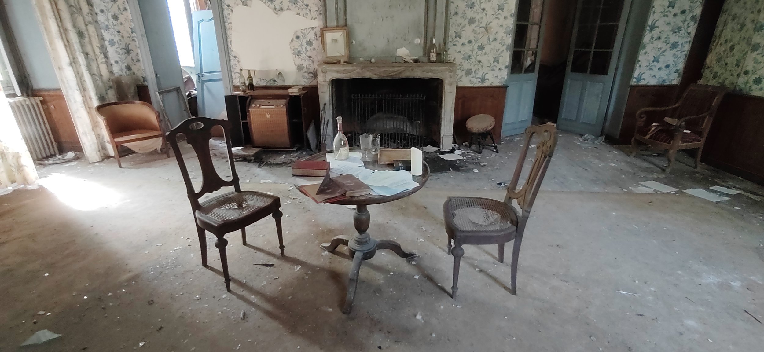 Ein verlassener Raum mit Staub und Müll auf dem Boden, alte Möbel, darunter ein runder Tisch mit Büchern, Papieren, einer Flasche, Kerze und Glas, sowie Stühle, ein Kamin, und verschlossene Türen und Fenster.