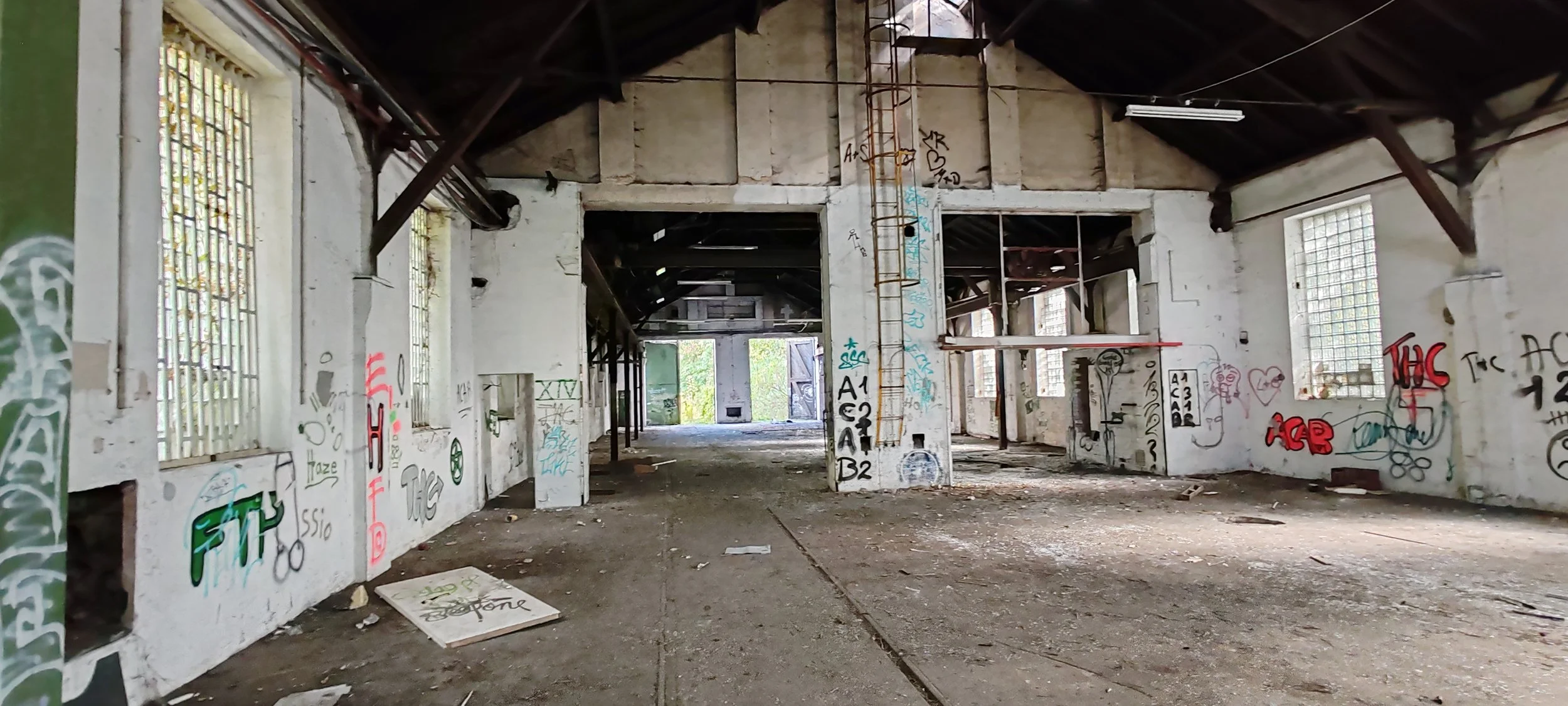 Verlassene, graffiti-bedeckte Industriehalle mit offenen Türen und Fenstern, Staub und Schmutz auf dem Boden, sichtbar durch zerbrochene Fenster und offene Türen