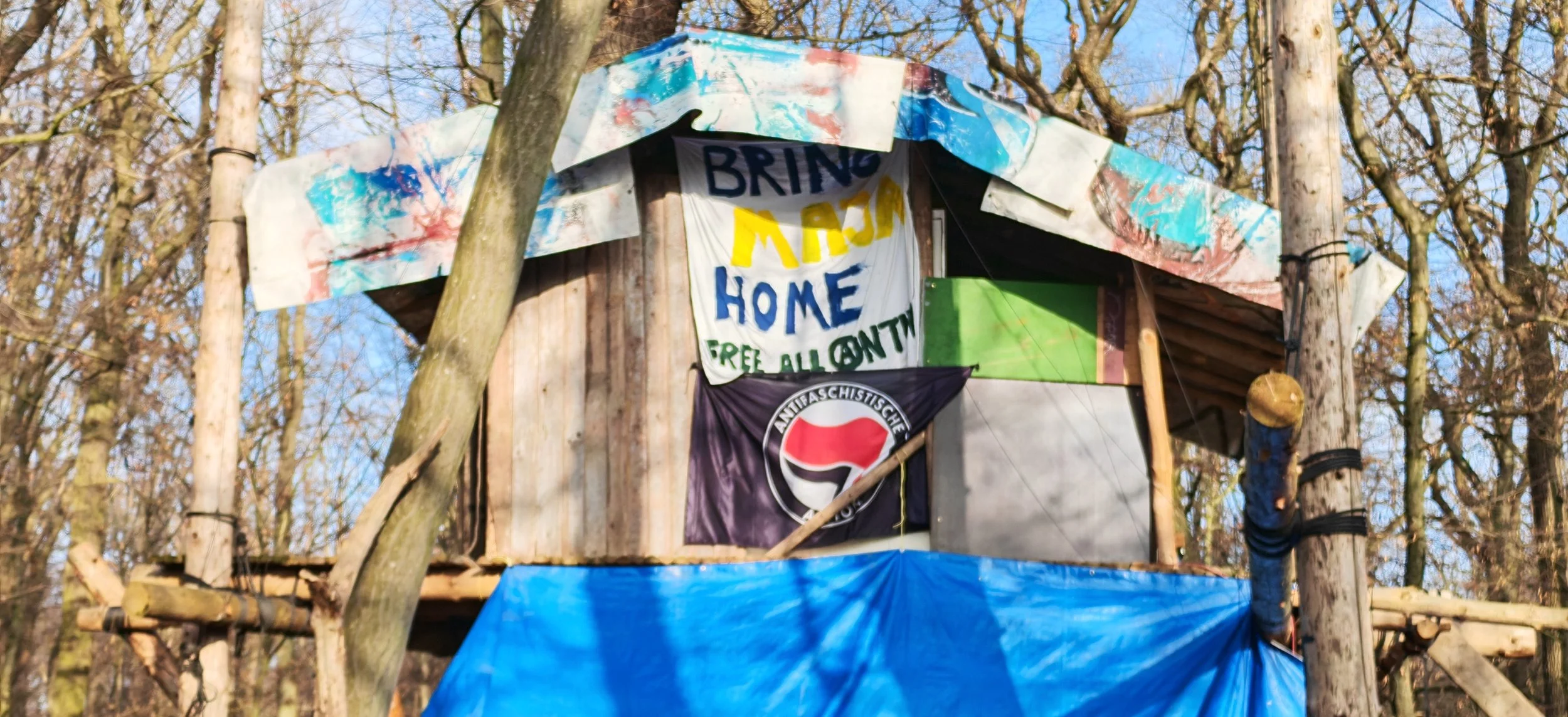 Eine Baumhütte im Wald mit mehreren Fahnen und einem Banner, das 'Bring Me Home Free All Continents' sagt.
