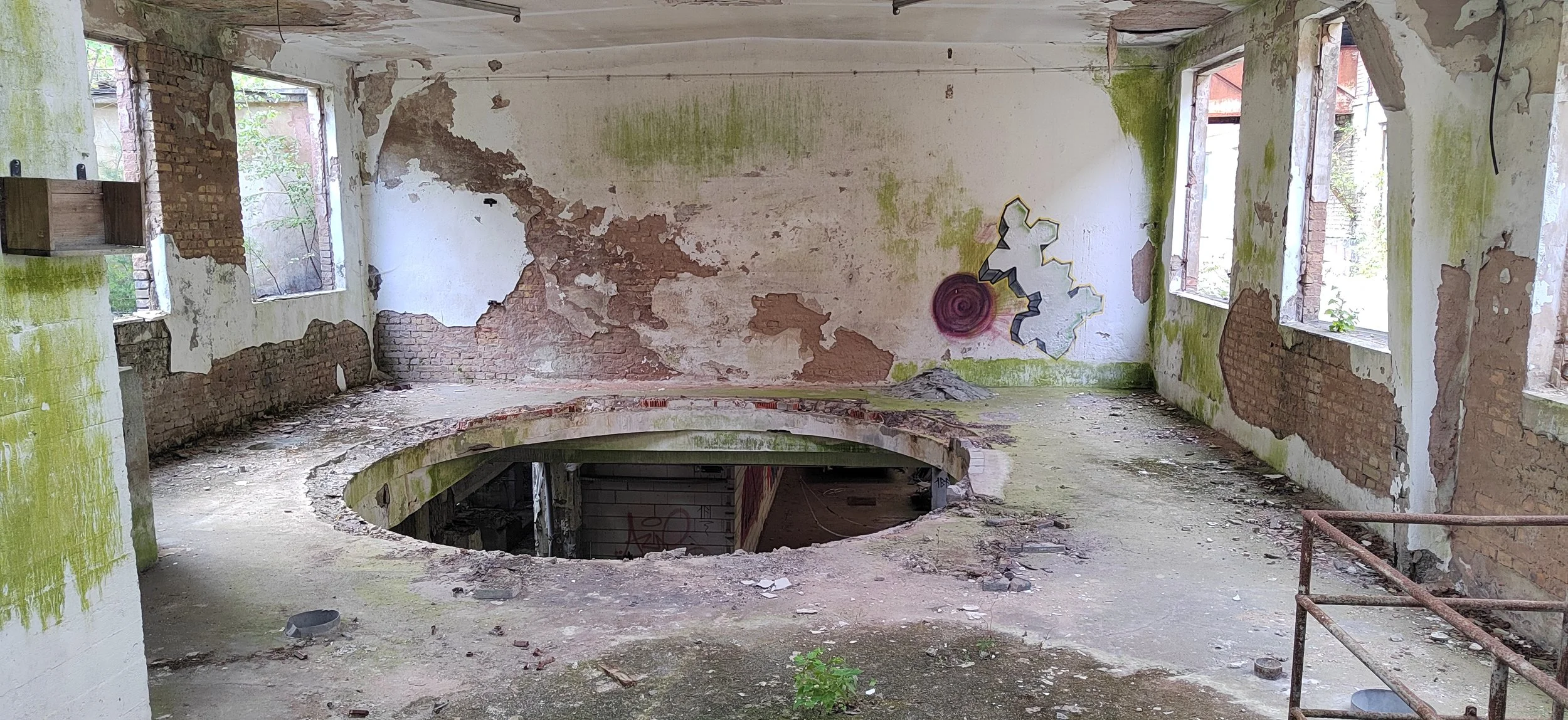 Verlassener, verfallener Raum mit zerbrochenen Wänden, moosbedeckten Oberflächen und Graffiti auf einer Wand, große offene Fenster, die natürliches Licht hereinlassen, und ein Stahlgeländer im Vordergrund.