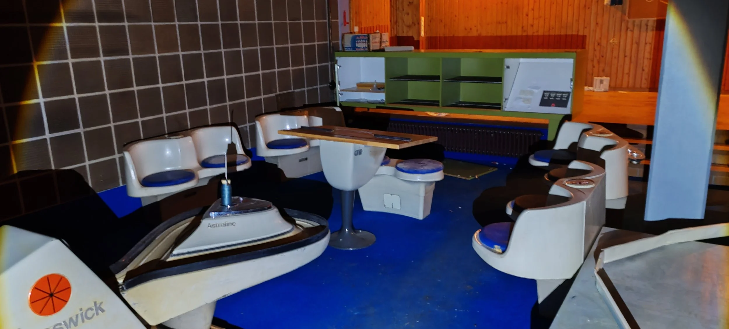 Sternenkreisel und Sitzgelegenheiten in einem Raum mit Holzverkleidung und blauer Fußboden. Es gibt eine Maschine mit dem Label "Astrofink" und eine grüne Schatztruhe im Hintergrund.