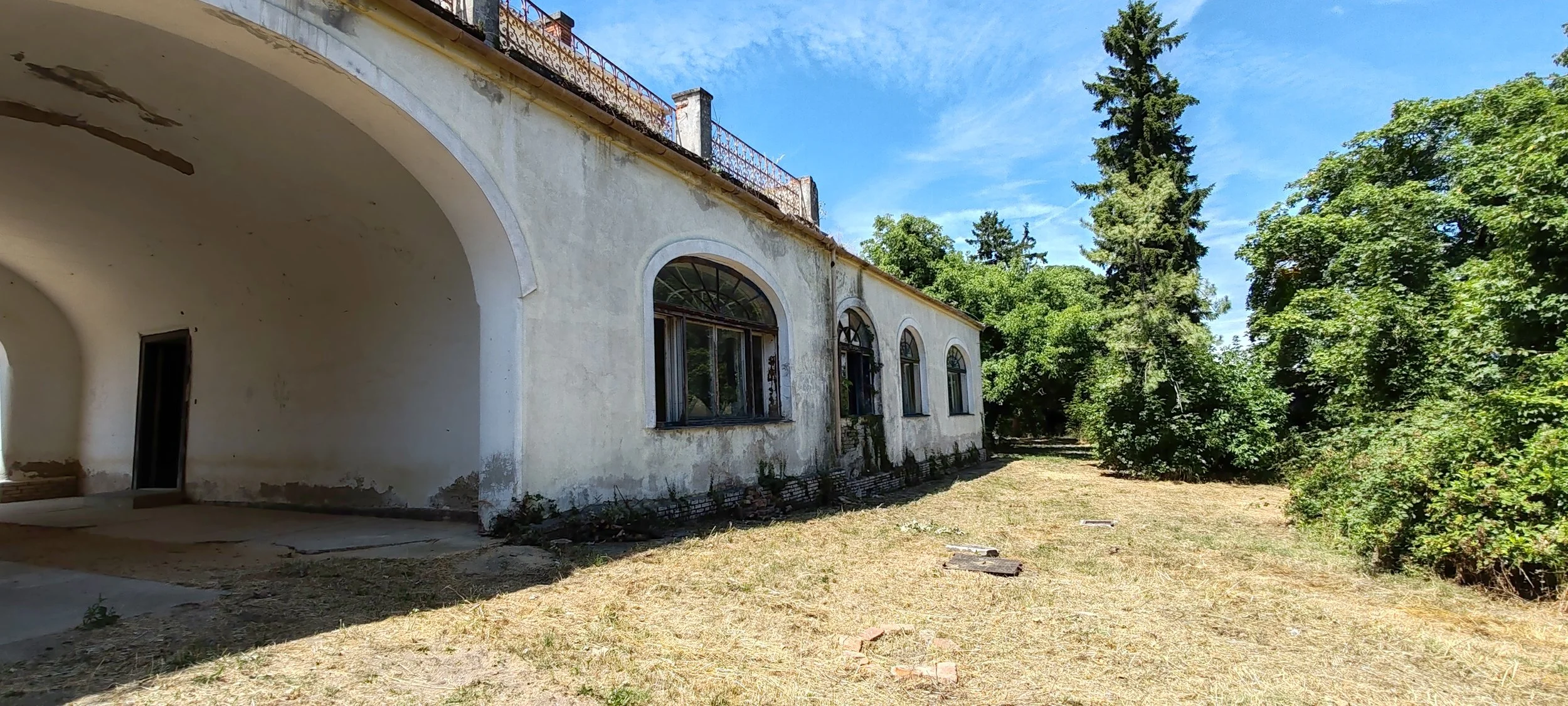 Ein altes, verfallenes Gebäude mit weißen, schmutzigen Wänden und abgenutzten Fenstern, umgeben von einer trockenen, kahlen Rasenfläche und vielen grünen Bäumen bei sonnigem Himmel.