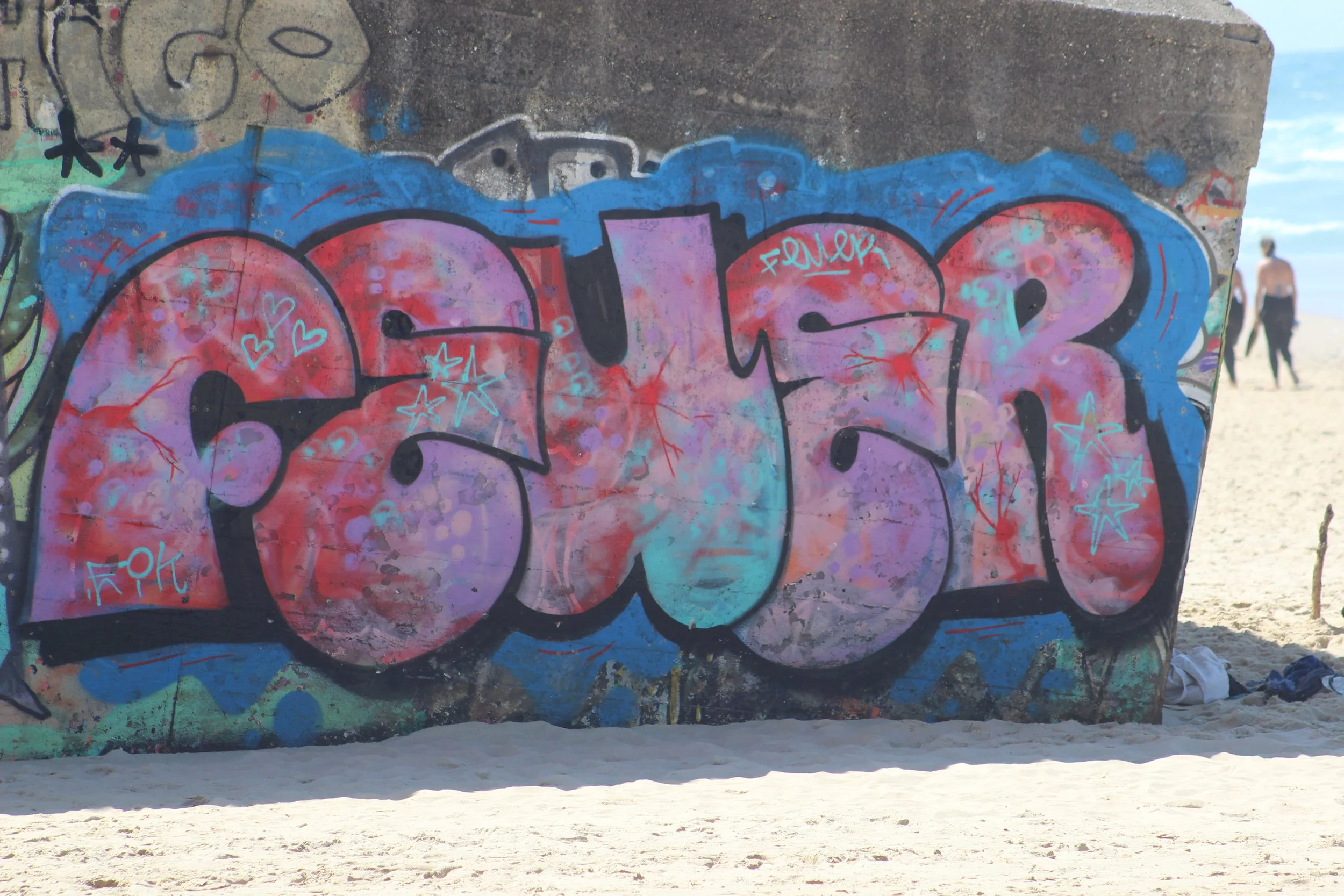 Bunte Graffiti auf einer Betonmauer am Strand mit Sand und strandende Menschen im Hintergrund.