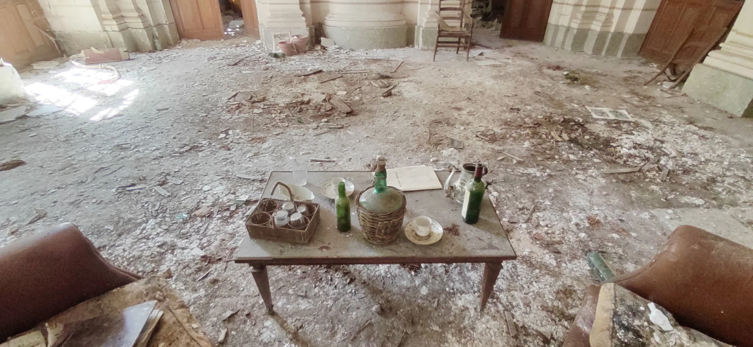 ein zerbrochenes und vernachlässigtes Interieur eines Raums mit Staub und Schutt, eine alte Tisch mit Getränkeflaschen, Geschirr und Papieren, umgeben von abgenutzten Sesseln und beschädigten Wänden.