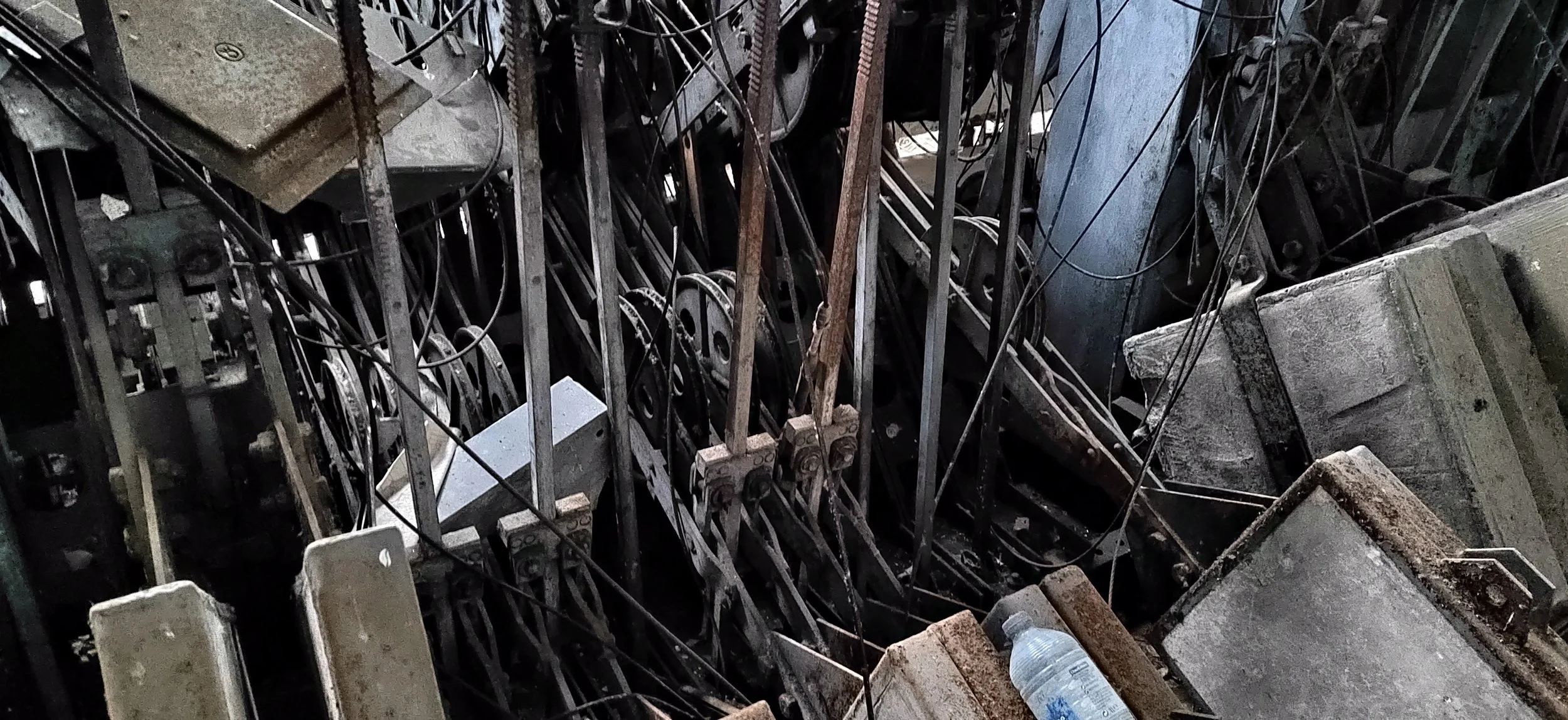 Ein chaotischer Haufen aus alten, rostigen Maschinenteilen, Drähten und Metallplatten in einem Schuppen oder einer Werkstatt, mit einer Wasserflasche im Vordergrund.