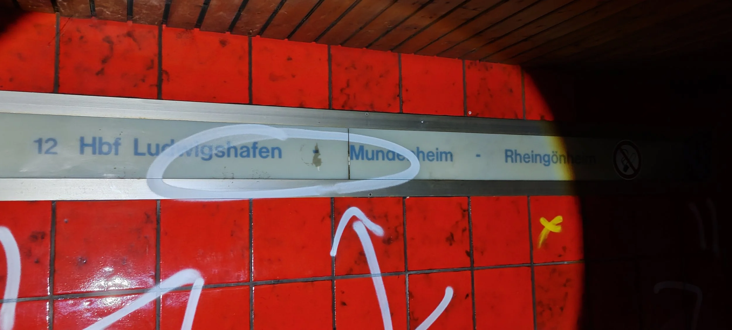 Der Name eines Bahnhofs im öffentlichen Nahverkehr, vermutlich in Deutschland, auf einem Schild mit roten Fliesen im Hintergrund, das mit weißer Farbe übermalt wurde.