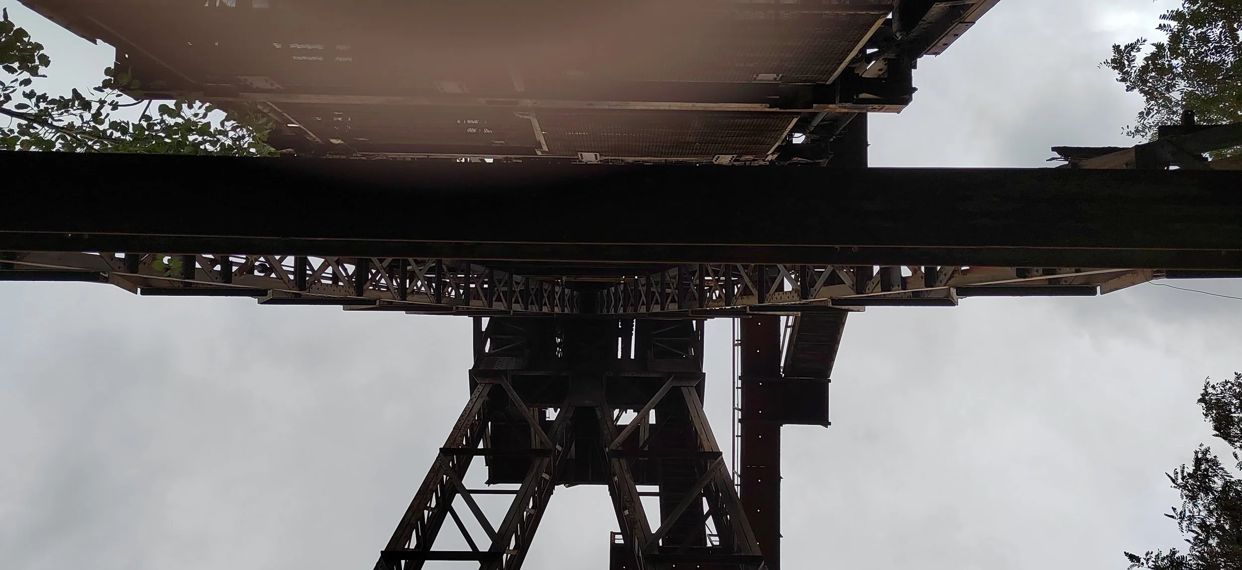 Blick von unten auf eine alte Eisenbahnbrücke mit Wolken am Himmel und einigen Bäumen im Hintergrund.