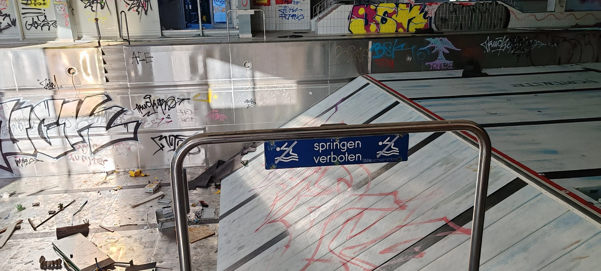 Ein verlassener und verwüsteter Skatepark mit Graffiti an den Wänden. Es liegt Müll und Schrott auf dem Boden. Ein Schild am Geländer verbietet das Springen ins Wasser.