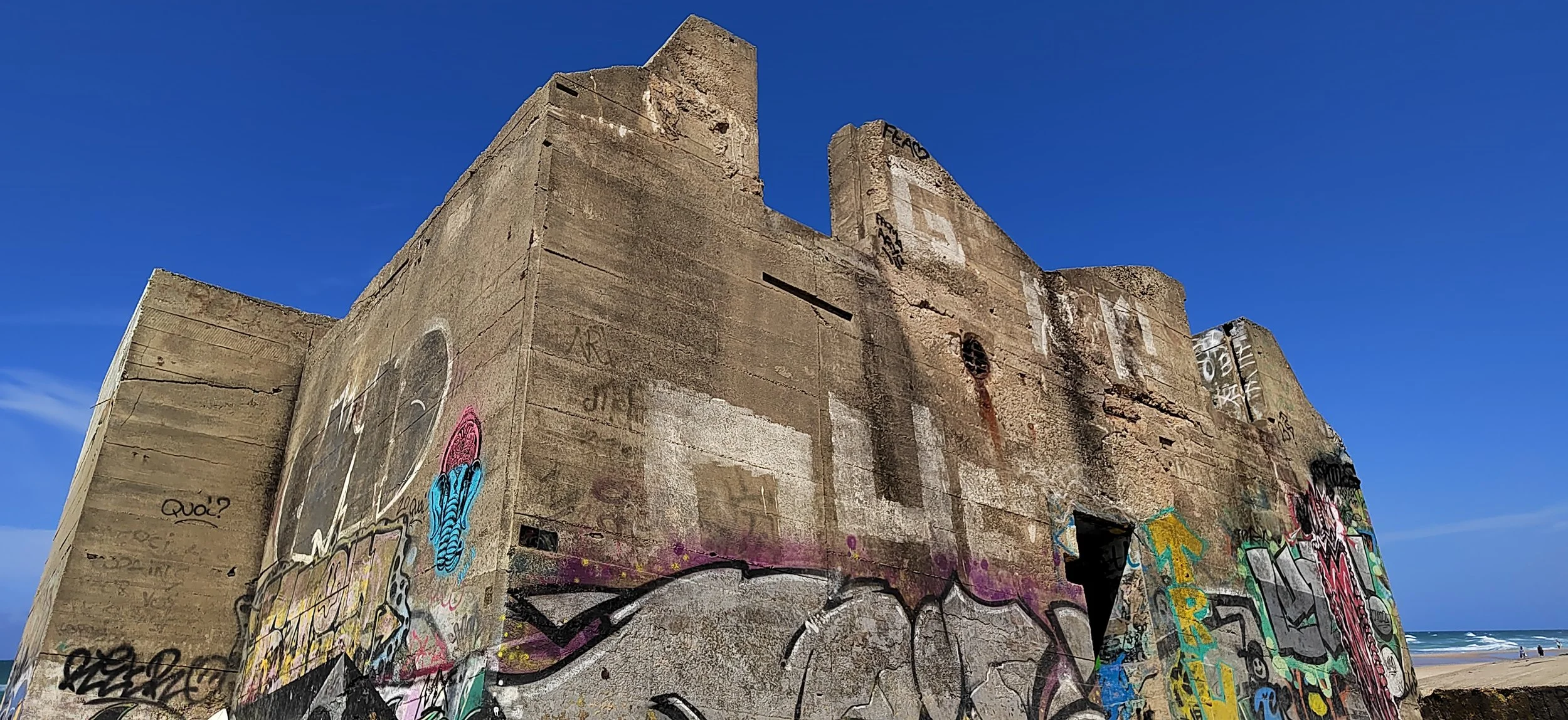 Verfallene Betonwand am Strand mit Graffiti, blauer Himmel im Hintergrund.
