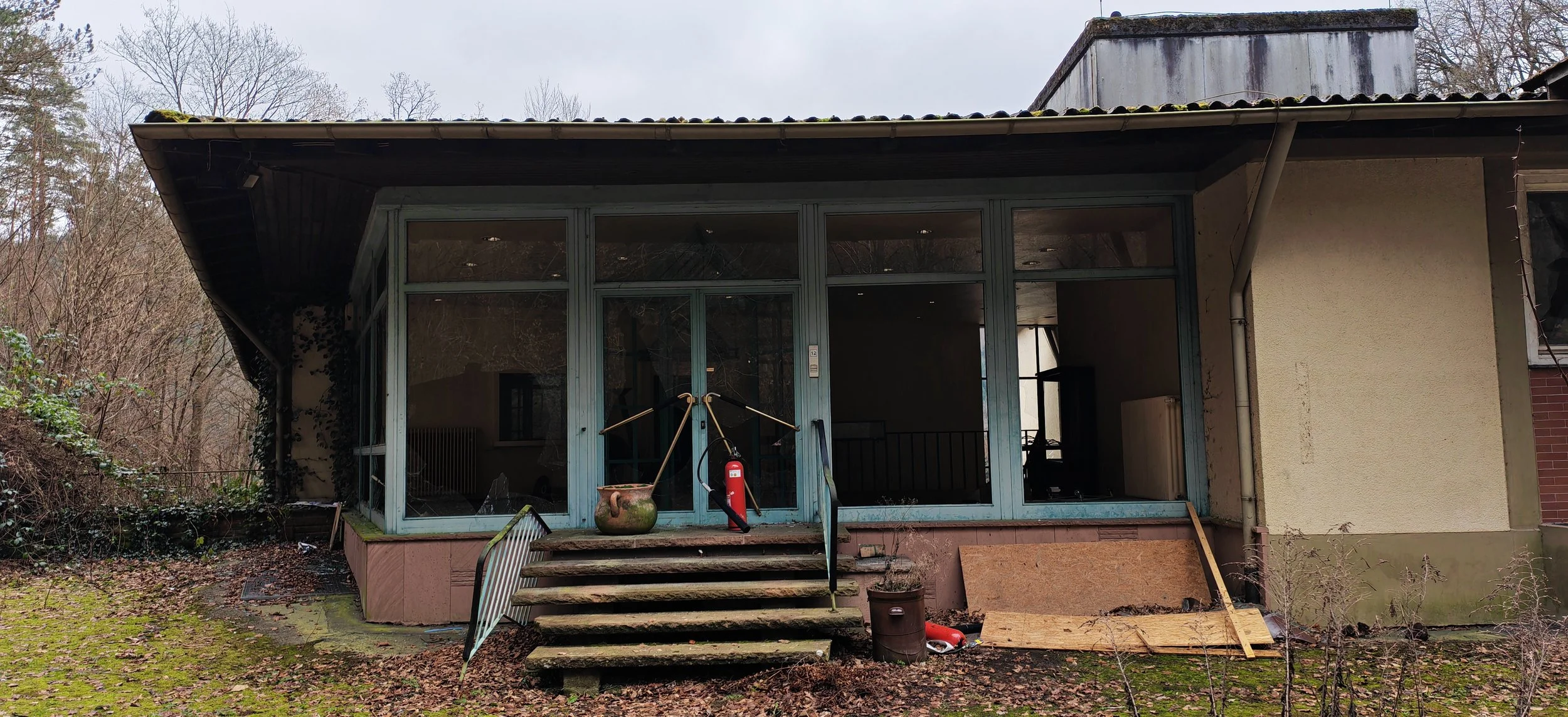 Verfallenes Haus mit breiten Stufen, Fenster und einer vorspringenden Überdachung, umgeben von Laub und Zweigen.