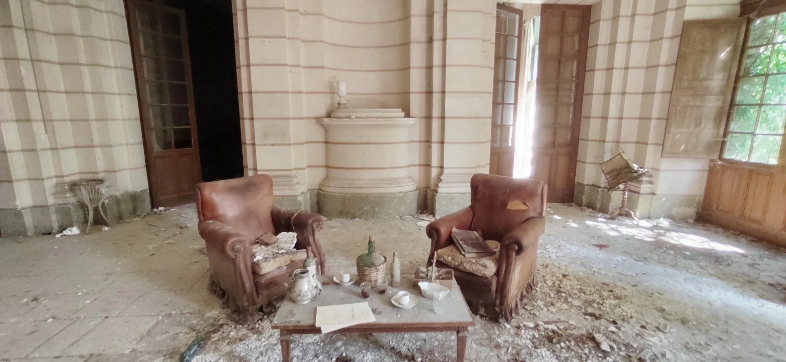 Verfallene, verlassene Wohnzimmer mit zwei abgenutzten, braunen Sesseln, einem kleinen Tisch mit alten Gegenständen und zerbrochenen Gläsern, auf zementiertem Boden mit Staub und Schutt, umgeben von wackeligen Möbeln, alten Fenster und Holz-Türen.