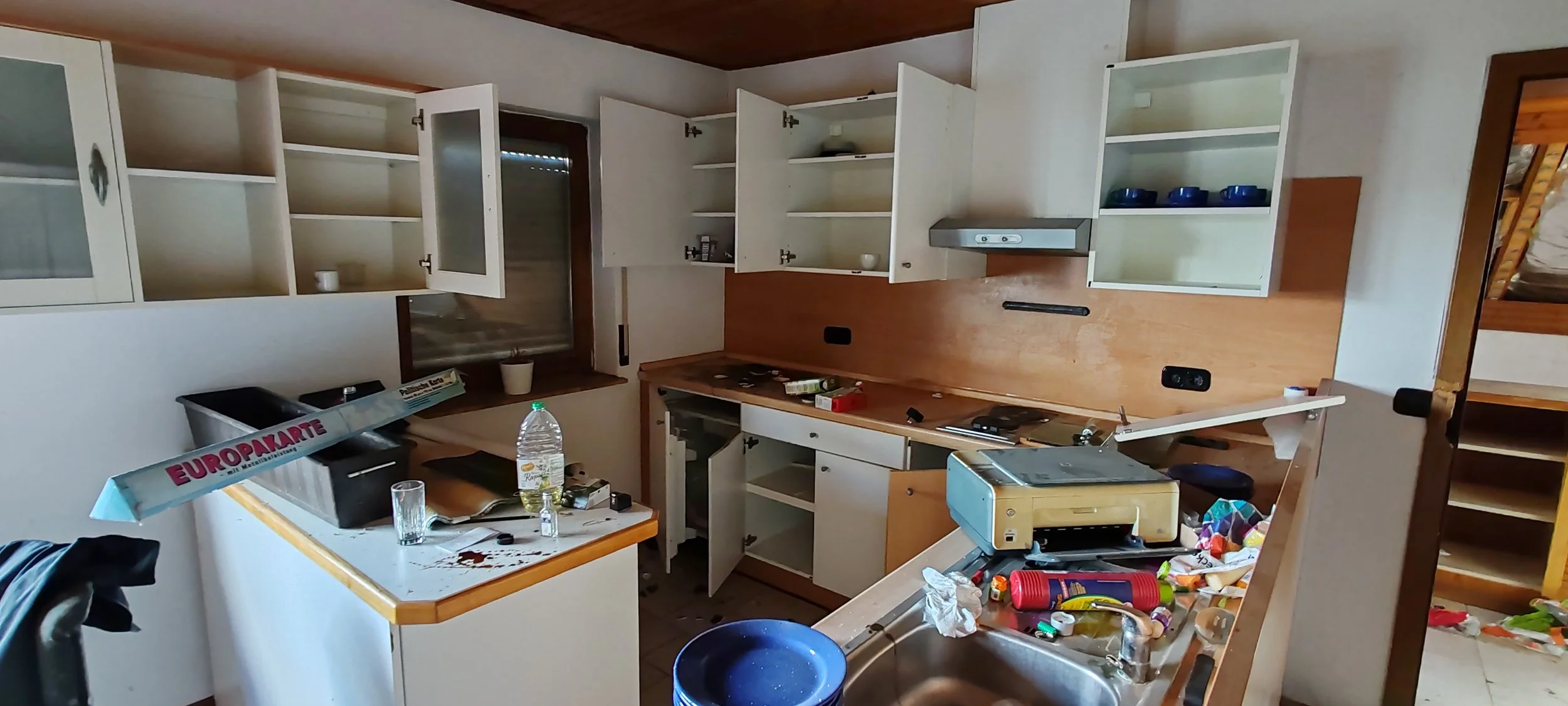 Un chaotisches, sauberes Küchenchaos mit offenen Schränken, unordentlichen Oberflächen und verstreuten Gegenständen wie Wasserflasche, Gläser, Zeitungen, Küchenutensilien, Verpackungen und Lebensmitteln.