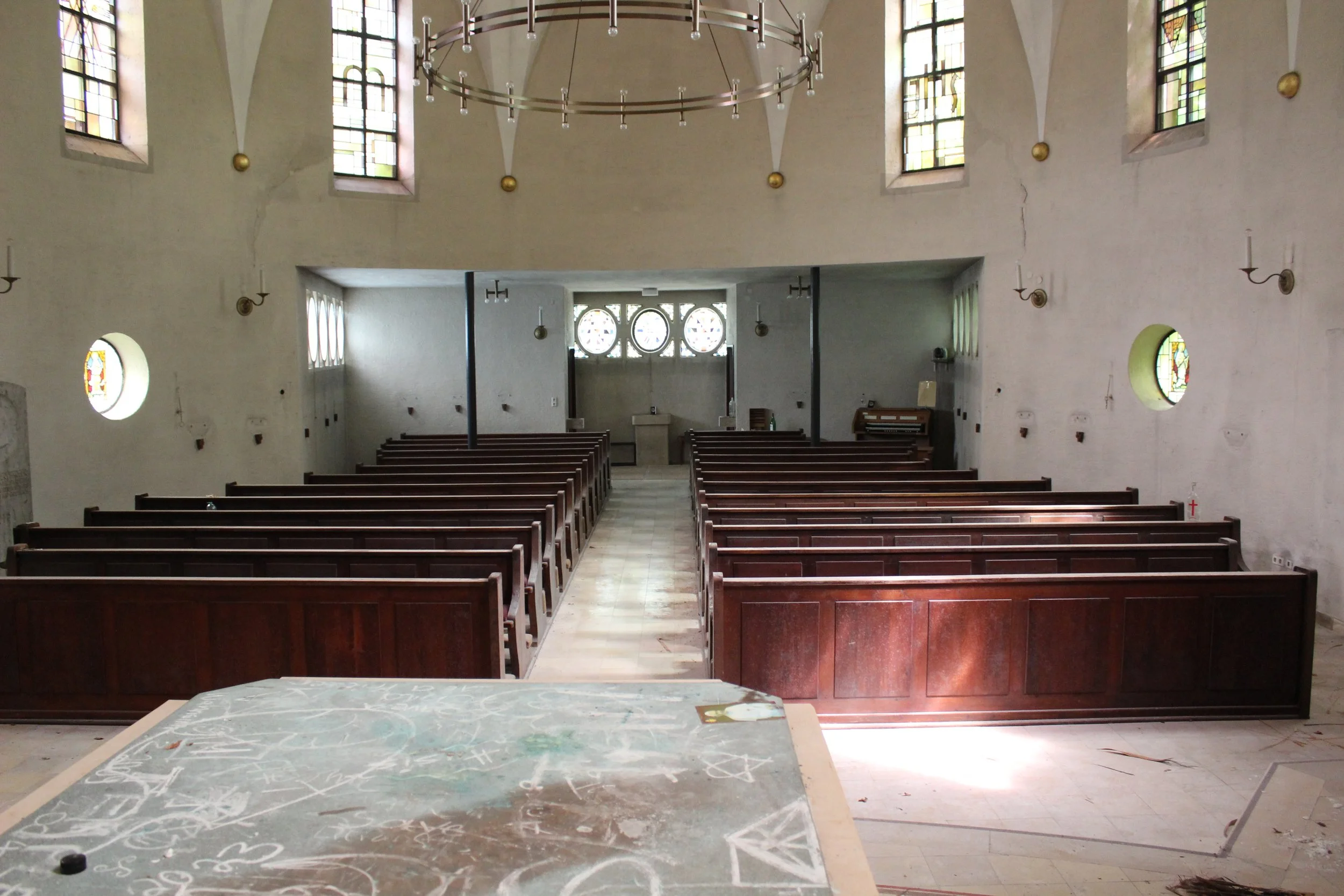 Innenraum einer leeren, alten Kirche mit Holzbänken, bunten Fenstern und einer großen Metall-Kabarettleuchter. Im Vordergrund ist eine beschmutzte Tafel zu sehen.