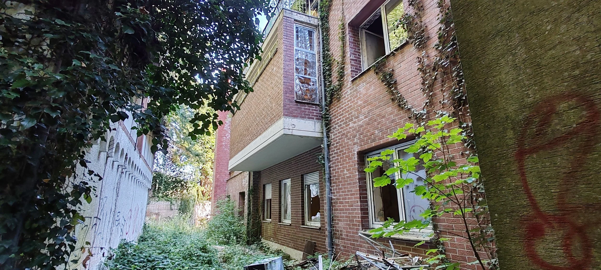 Stadthaus mit steinverzierten Fensterbalkonen, Fenster mit weißen Rollläden, verwitterte Mauer mit Graffiti, umgeben von Büschen und Bäumen, im Hintergrund sonniger Himmel.